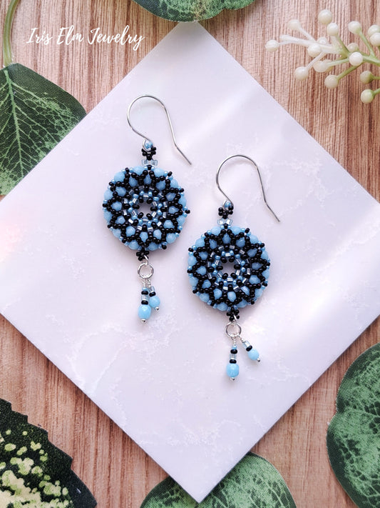 Pair of light blue and black glass beaded earrings on a white card with 'Iris Ella Jewelry' branding, placed on a wooden surface with green leaves.