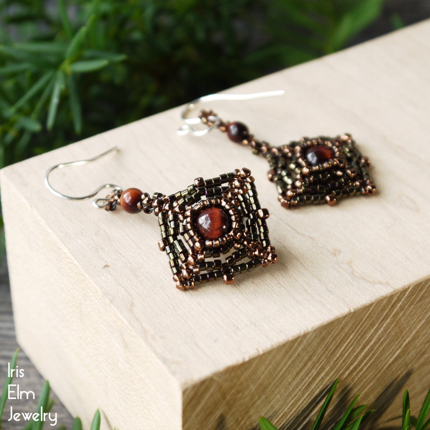Pair of handmade brown stone beaded red tiger eye earrings on a wooden block with greenery in the background, featuring the brand 'Iris Elm Jewelry'.