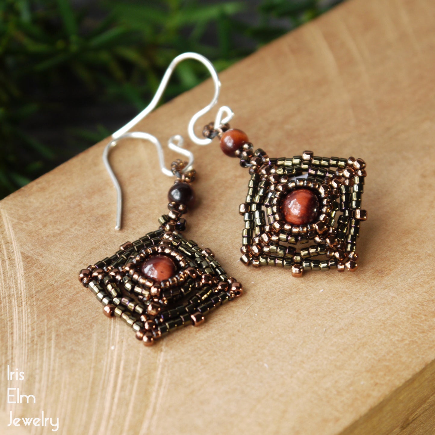 Pair of brown beaded square earrings with red tiger eye stones on a wooden surface, branded 'Iris Elm Jewelry'.