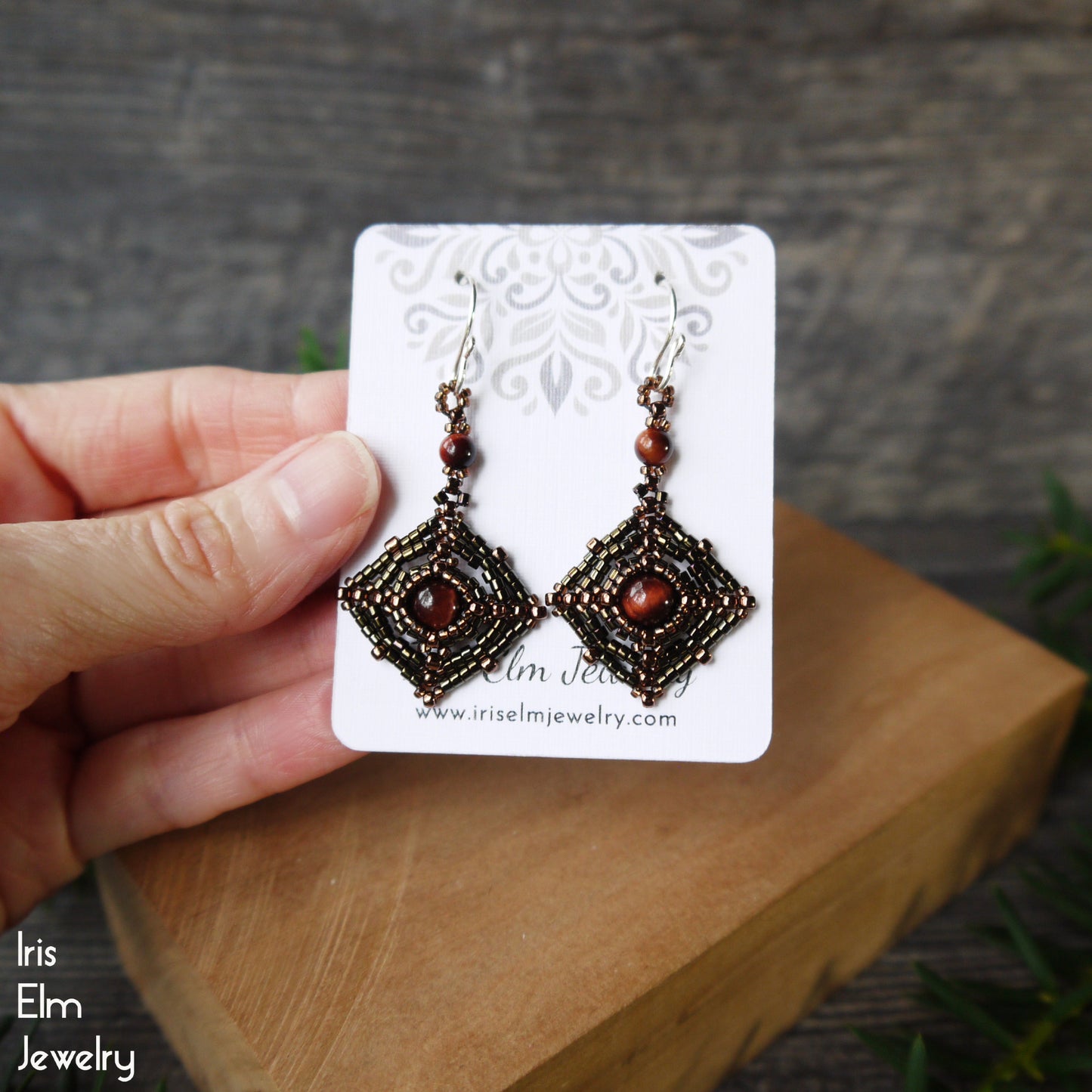 Pair of artisan beaded red tiger eye gemstone and seed bead earrings on a card held by a hand with a wooden block and grass background