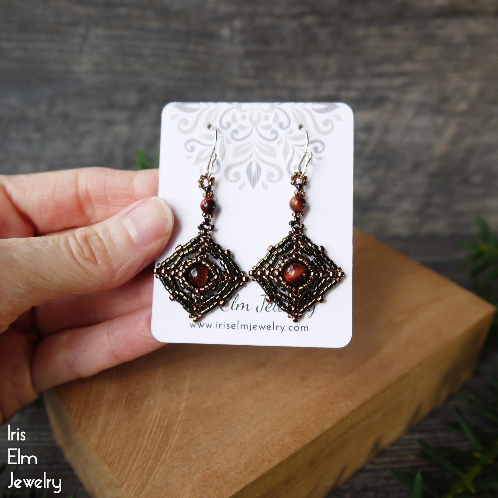 Pair of artisan beaded red tiger eye gemstone and seed bead earrings on a card held by a hand with a wooden block and grass background