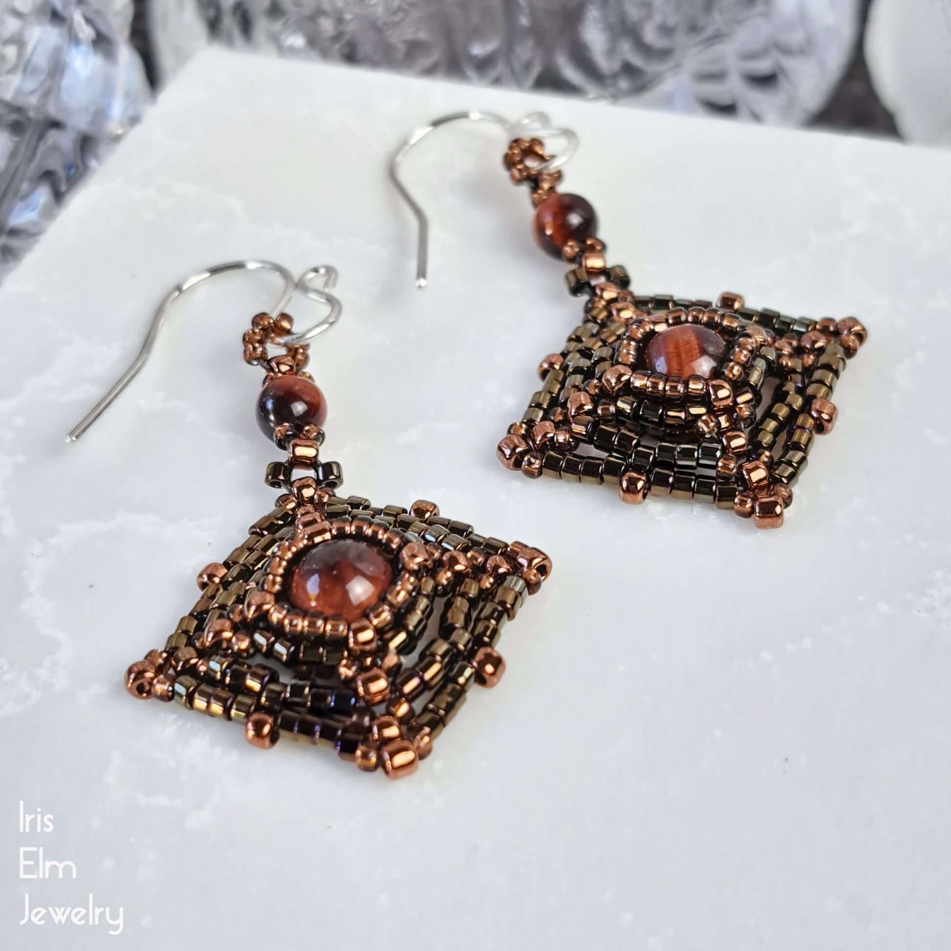 Pair of handmade beaded square earrings with brown gemstones in center on a white surface, branded 'Iris Elm Jewelry'.