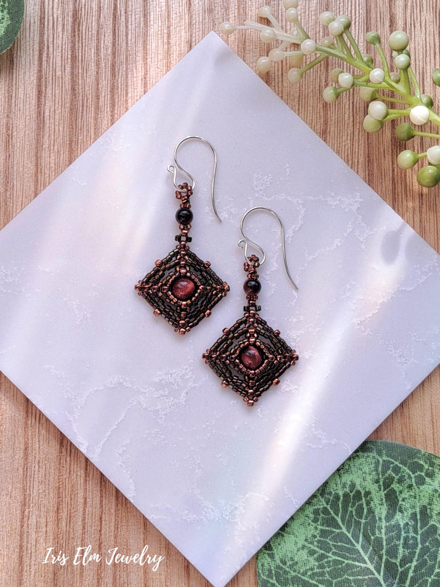 Pair of ornate beadwork red tiger eye earrings on a white tile with 'Iris Elm Jewelry' branding.