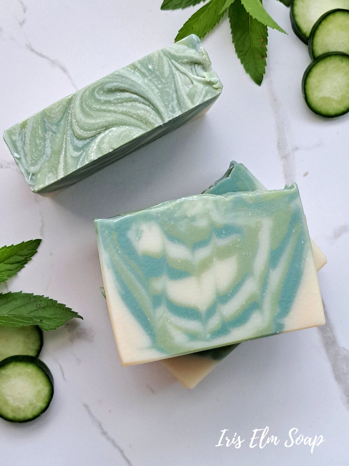 Two green and white swirled garden mint and cucumber handmade soap bars with cucumber slices and leaves on a light background, featuring 'Iris Elm Soap'.