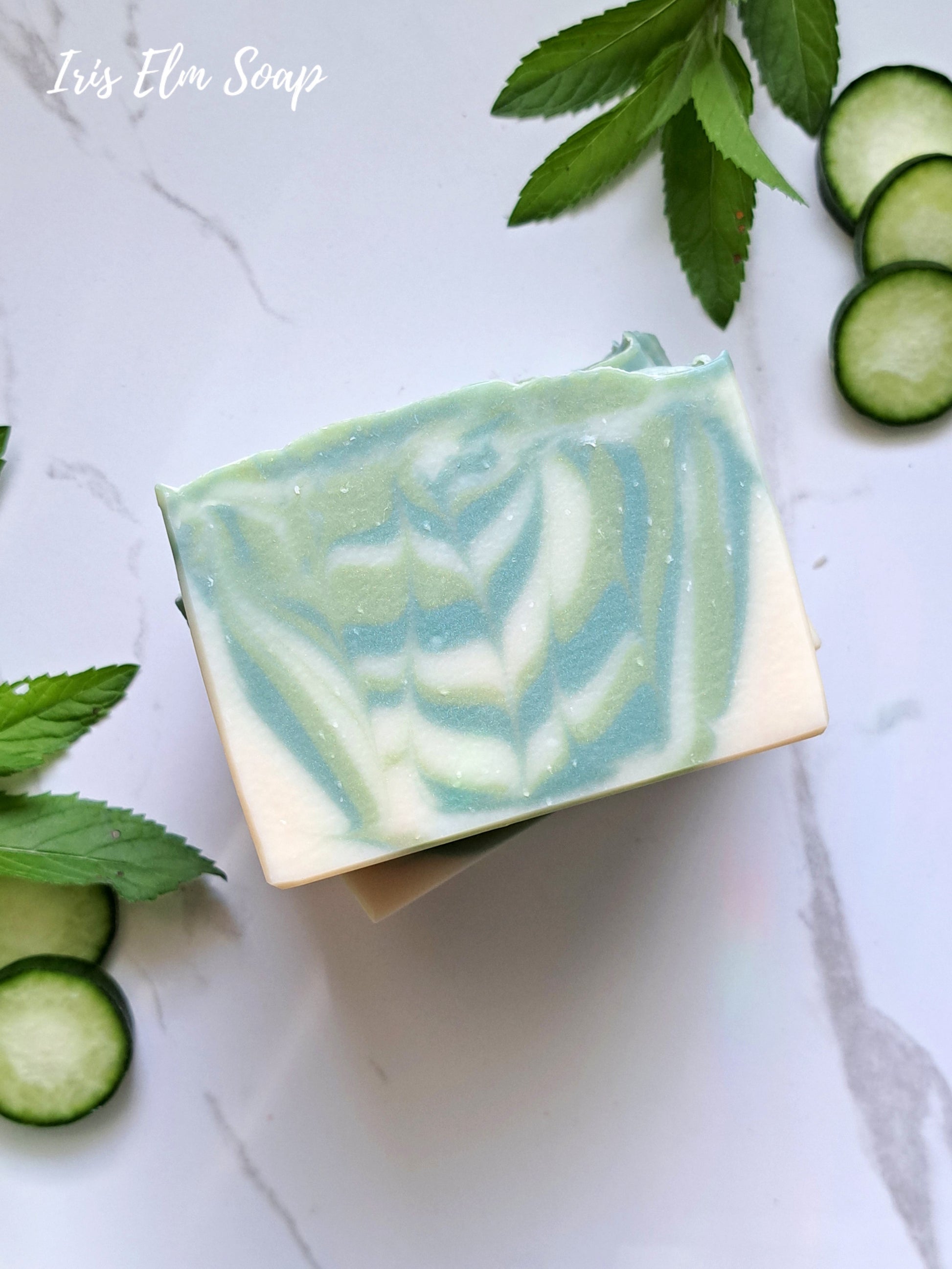 Bar of garden mint and cucumber handmade soap with cucumber slices and leaves on a marble surface, branded 'Iris Elm Soap'.