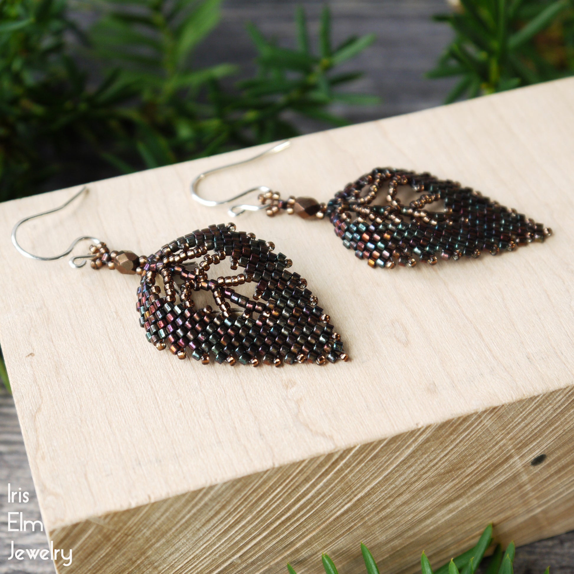 dark brown bronze handmade Leaf-shaped beaded seed bead earrings on a wooden block with greenery in the background