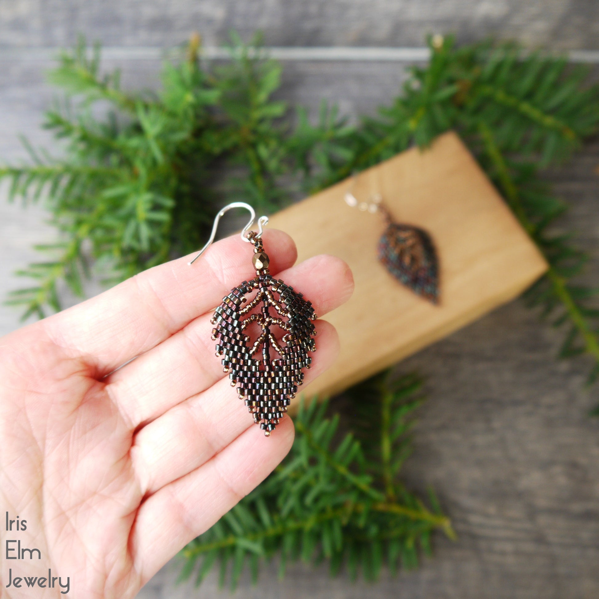 Hand holding a leaf-shaped seed bead woven earring with sterling silver ear wire with 'Iris Elm Jewelry' branding, against a natural background.