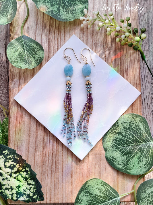 Tassel earrings with blue amazonite beads on a white card, surrounded by green leaves on a wooden surface.