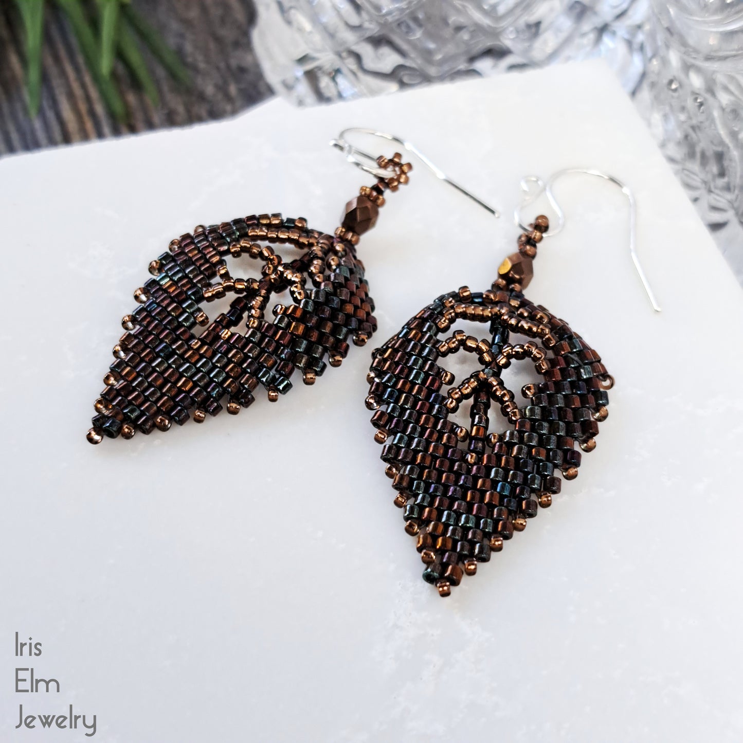 Beaded dark brown autumn leaf-shaped earrings on a white surface with 'Iris Elm Jewelry' branding.
