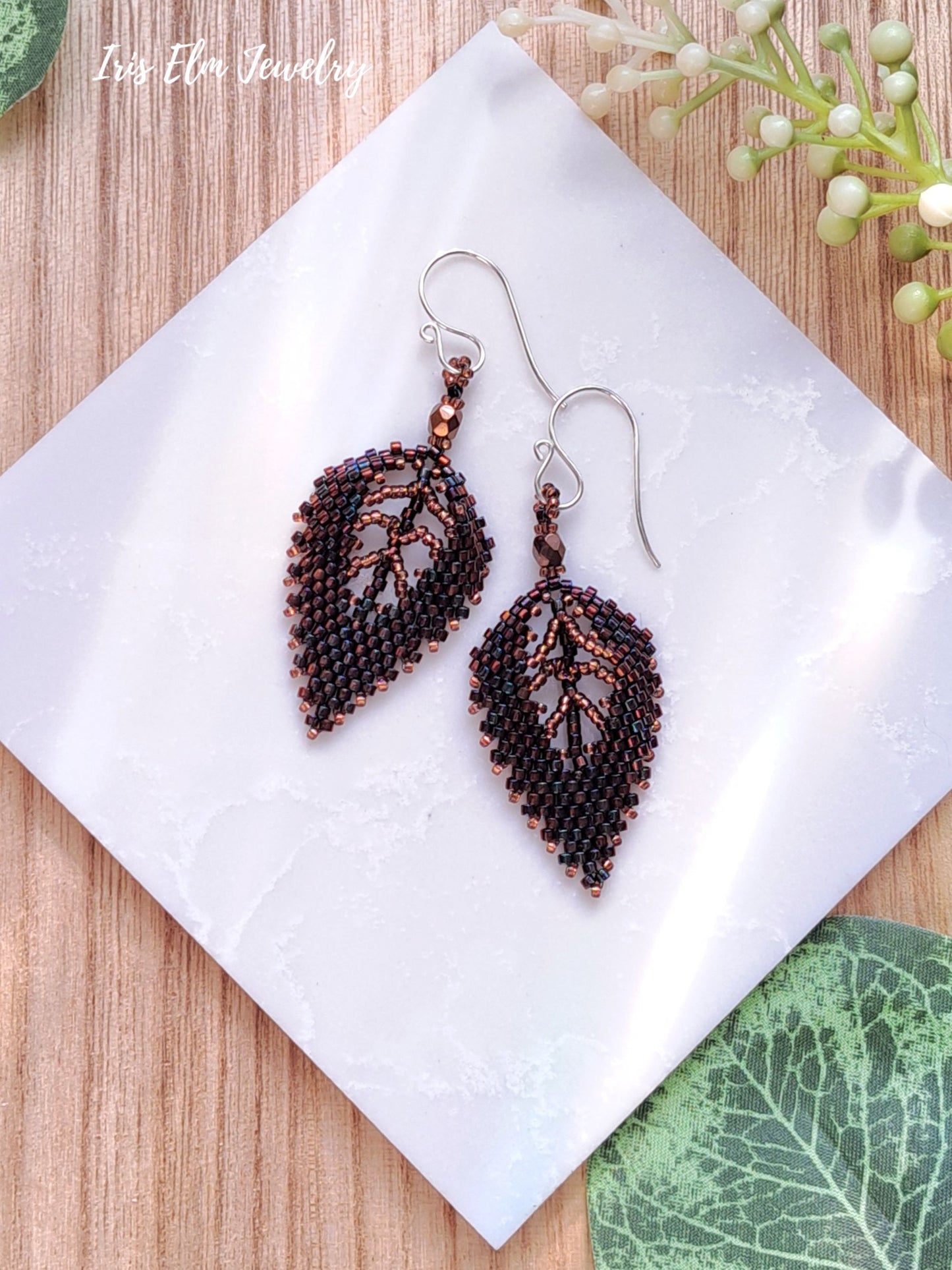 Pair of beaded autumn dark brown bronze seed bead leaf earrings on a white tile with green leaves in the background
