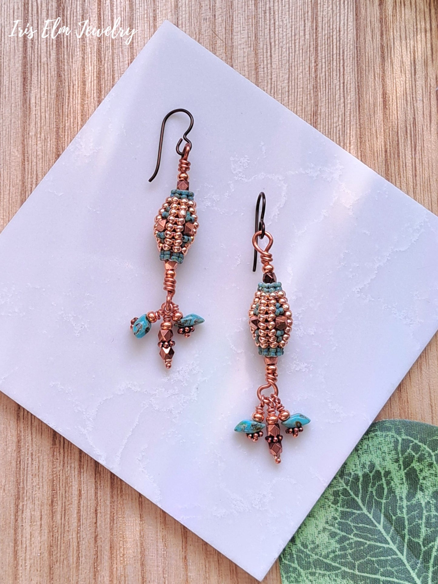 Pair of beaded copper and turquoise glass dangle earrings on a white square with a wooden background