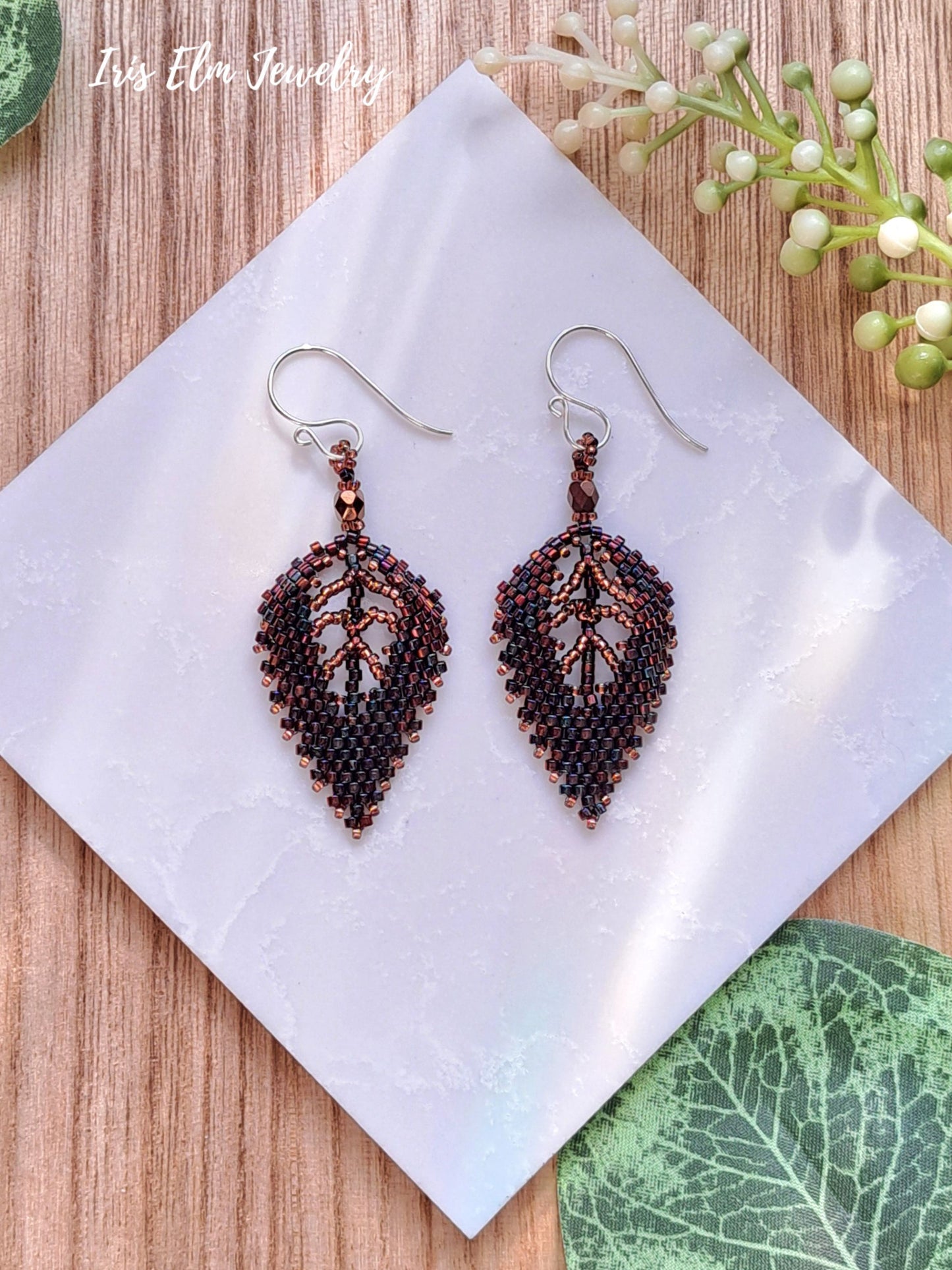 Leaf-shaped beaded dark brown seed bead earrings on a white napkin with green leaves in the background