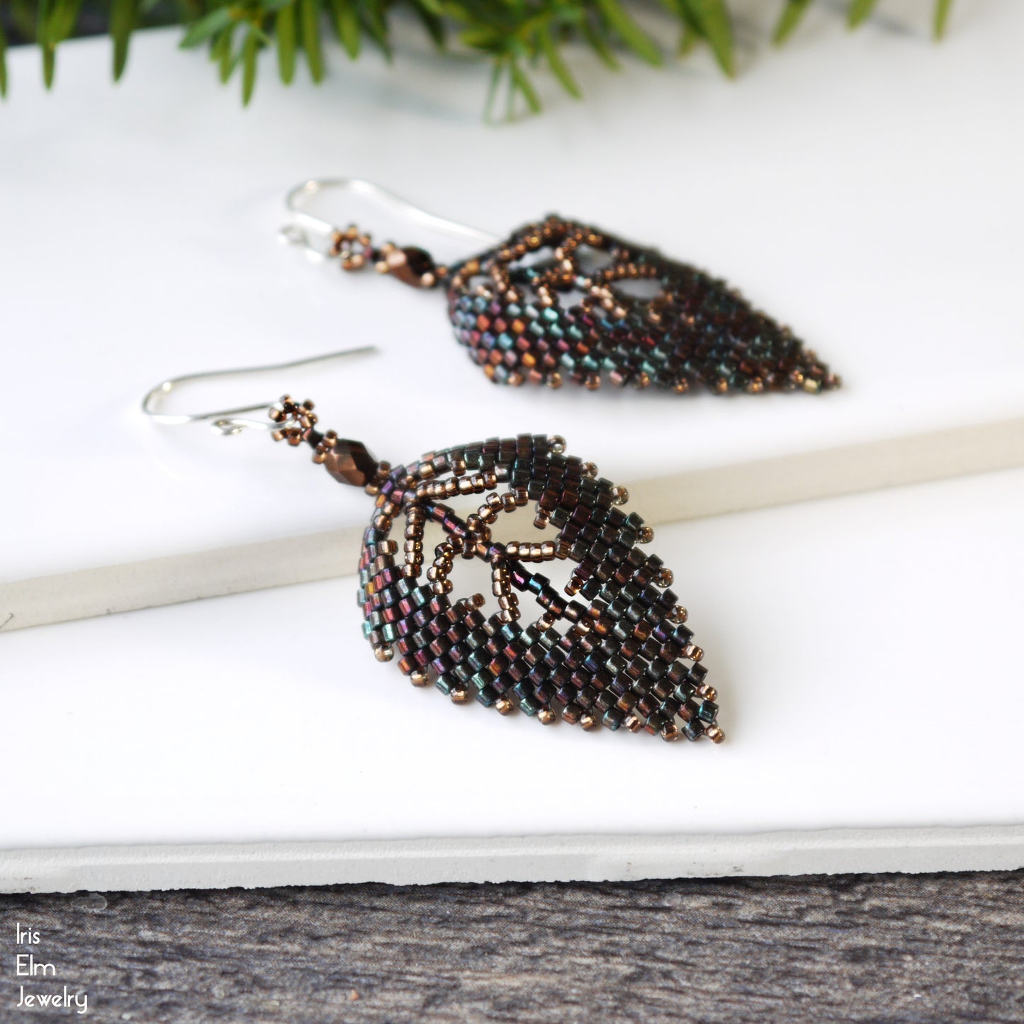 Leaf-shaped beaded brown glass handcrafted earrings on a white surface with greenery in the background