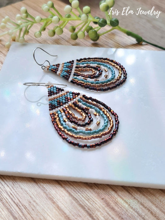 Pair of beaded seed bead bronze and aqua blue teardrop earrings on a white surface with a wooden background