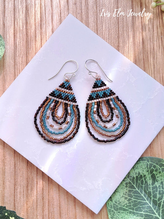 Pair of beaded seed bead teardrop bronze brown and aqua blue earrings on a white square with 'Iris Elm Jewelry' branding, placed on a wooden surface.