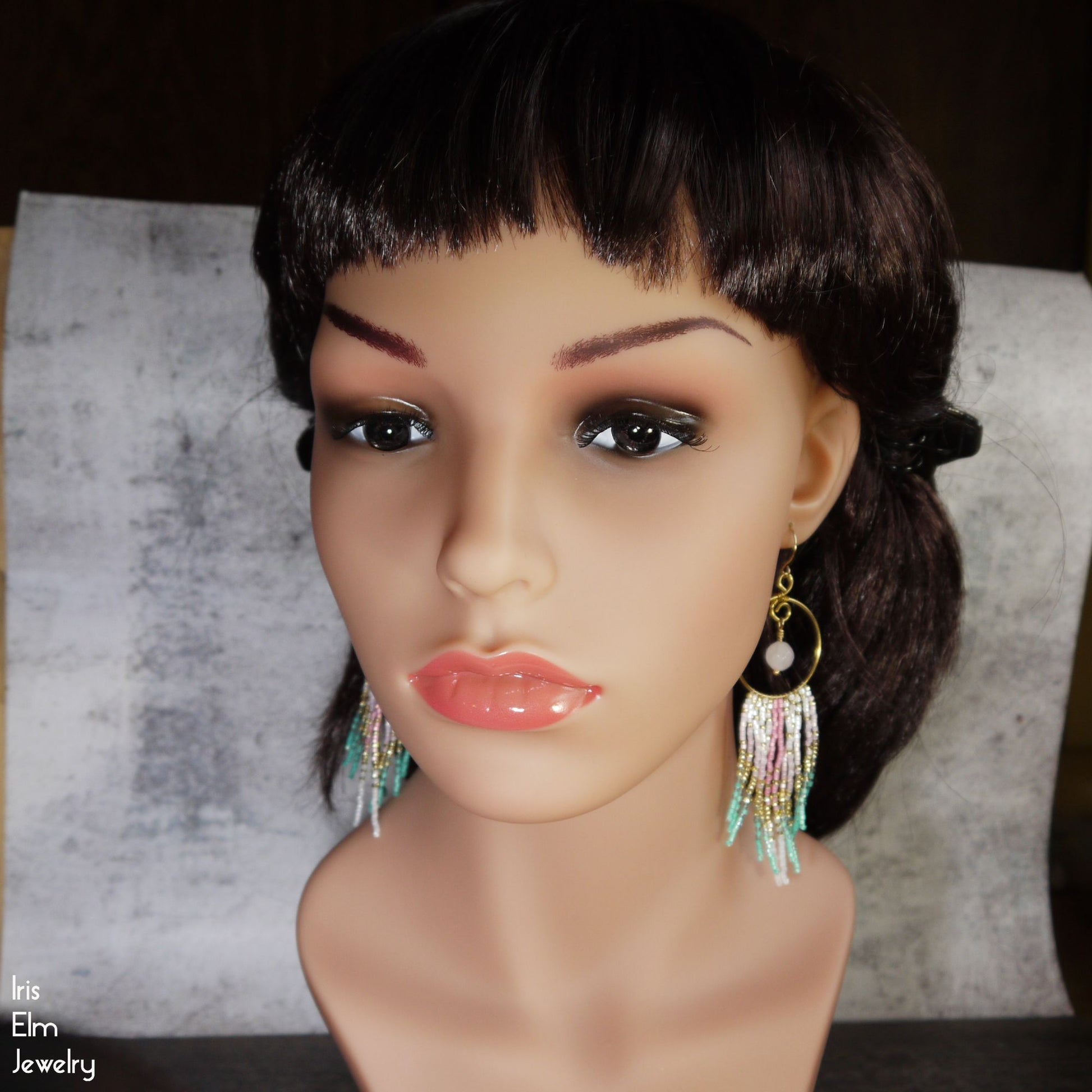 Mannequin head with black hair wearing colorful fringe earrings on a textured surface.