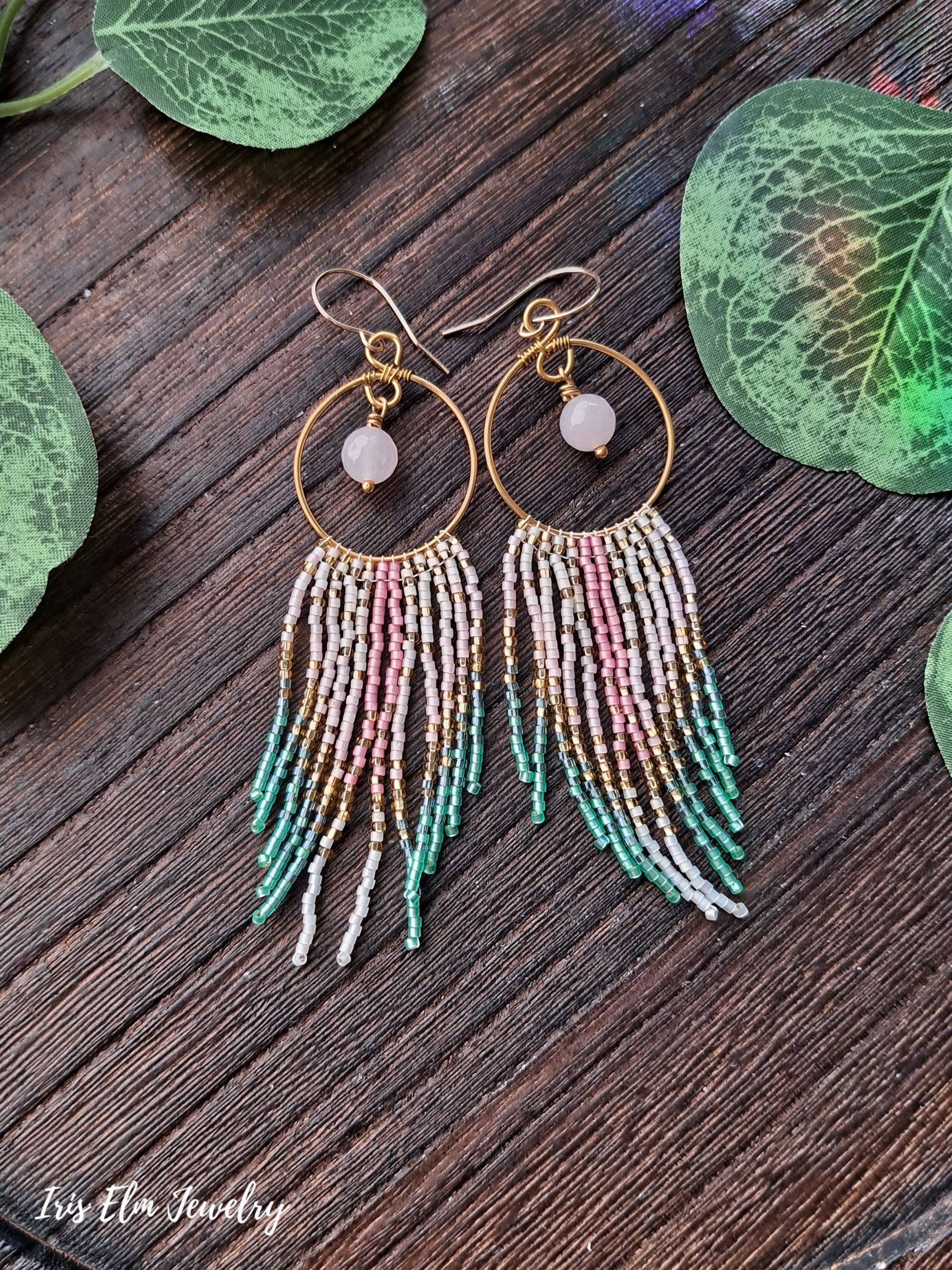 Gold hoop rose quartz earrings with colorful beaded fringe on a wooden surface with leaves.