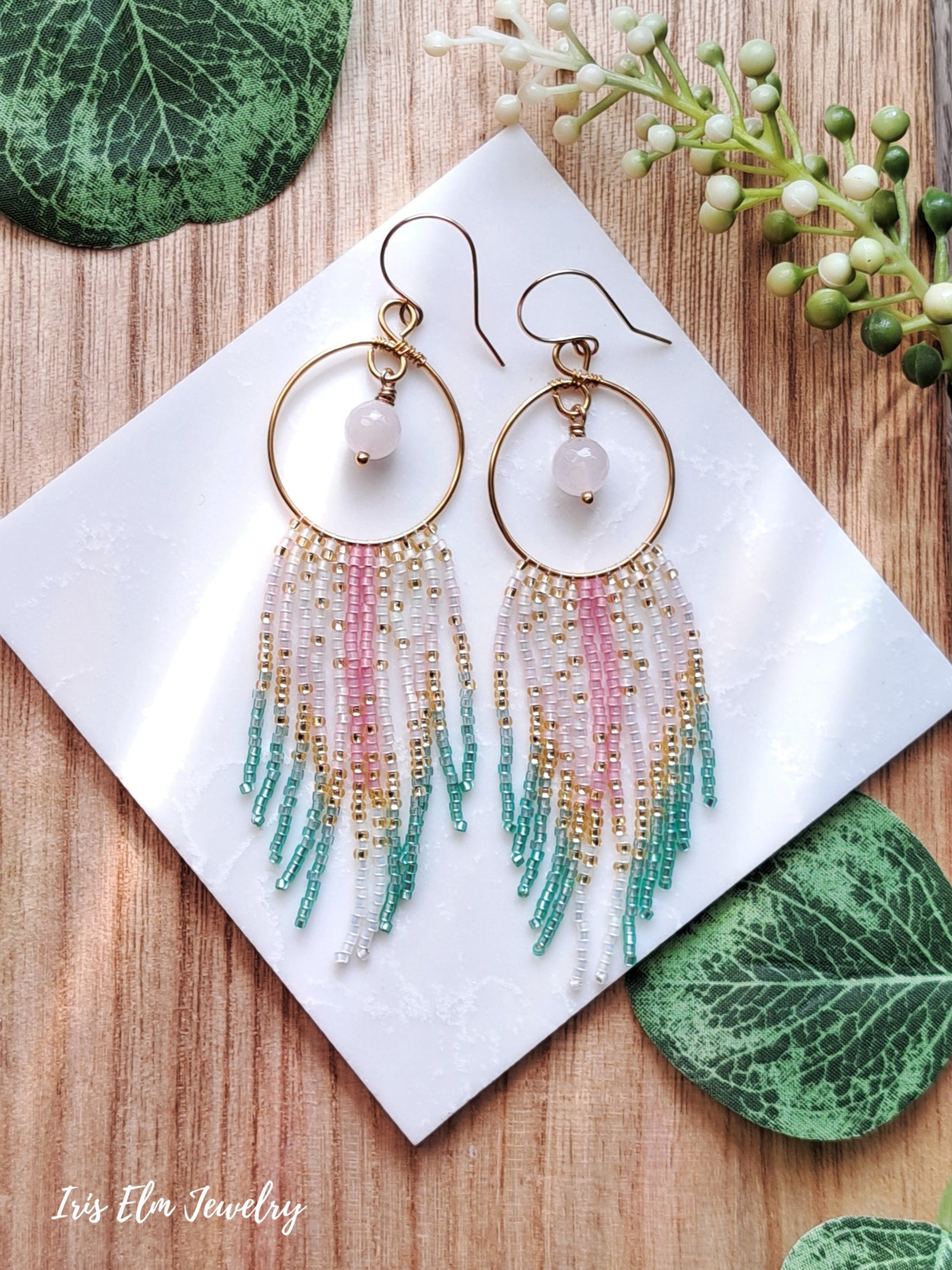 Multicolored beaded hoop fringe rose quartz earrings on a white card with green leaves in the background