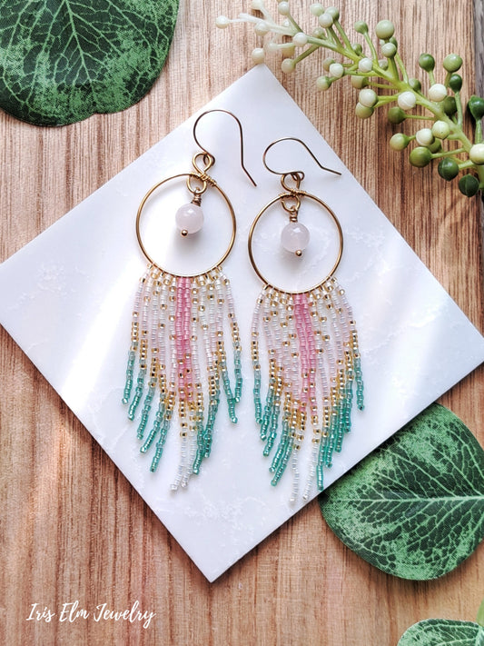 Multicolored beaded hoop fringe rose quartz earrings on a white card with green leaves in the background