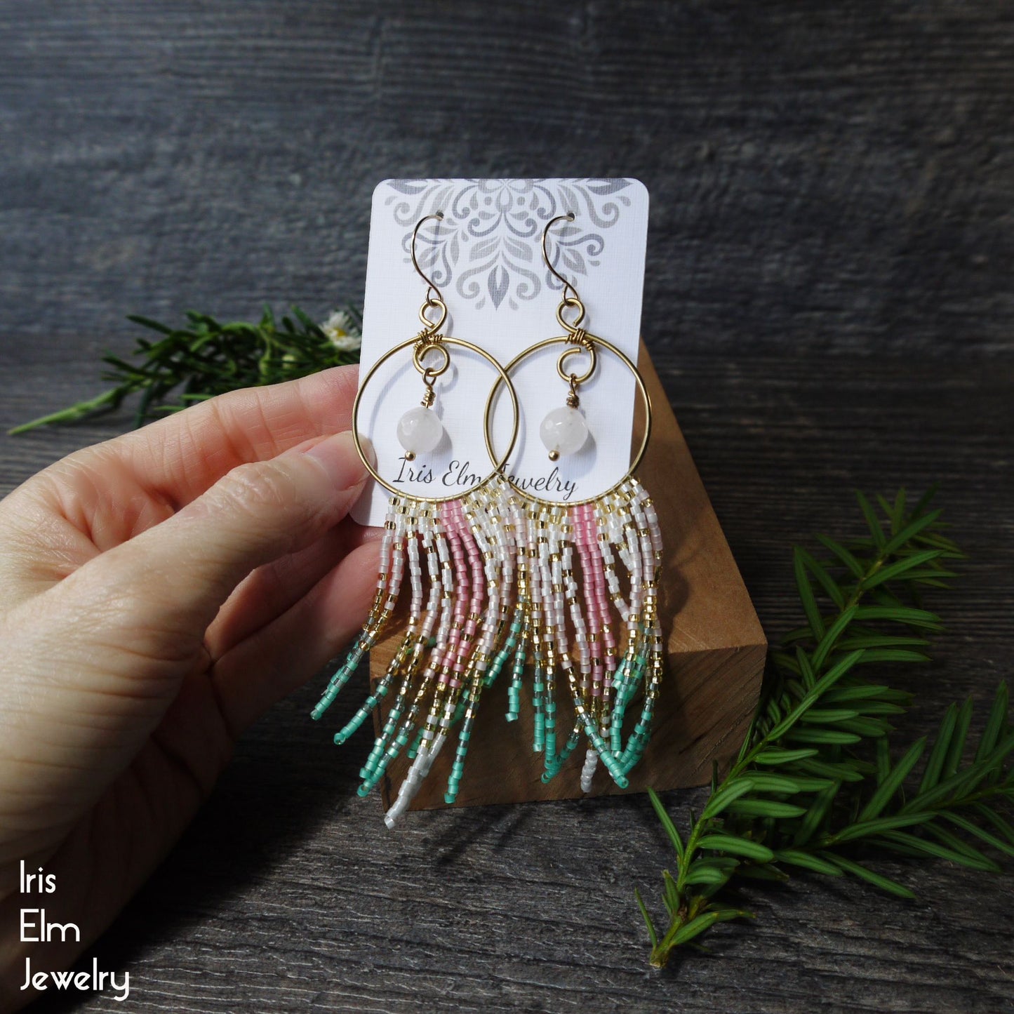 Hand holding a pair of beaded rose quartz fringe hoop earrings on a branded card with a dark wooden background.