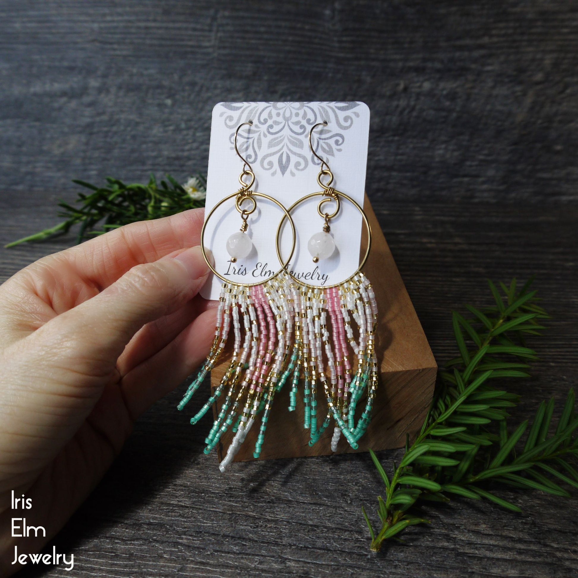 Hand holding a pair of beaded rose quartz fringe hoop earrings on a branded card with a dark wooden background.