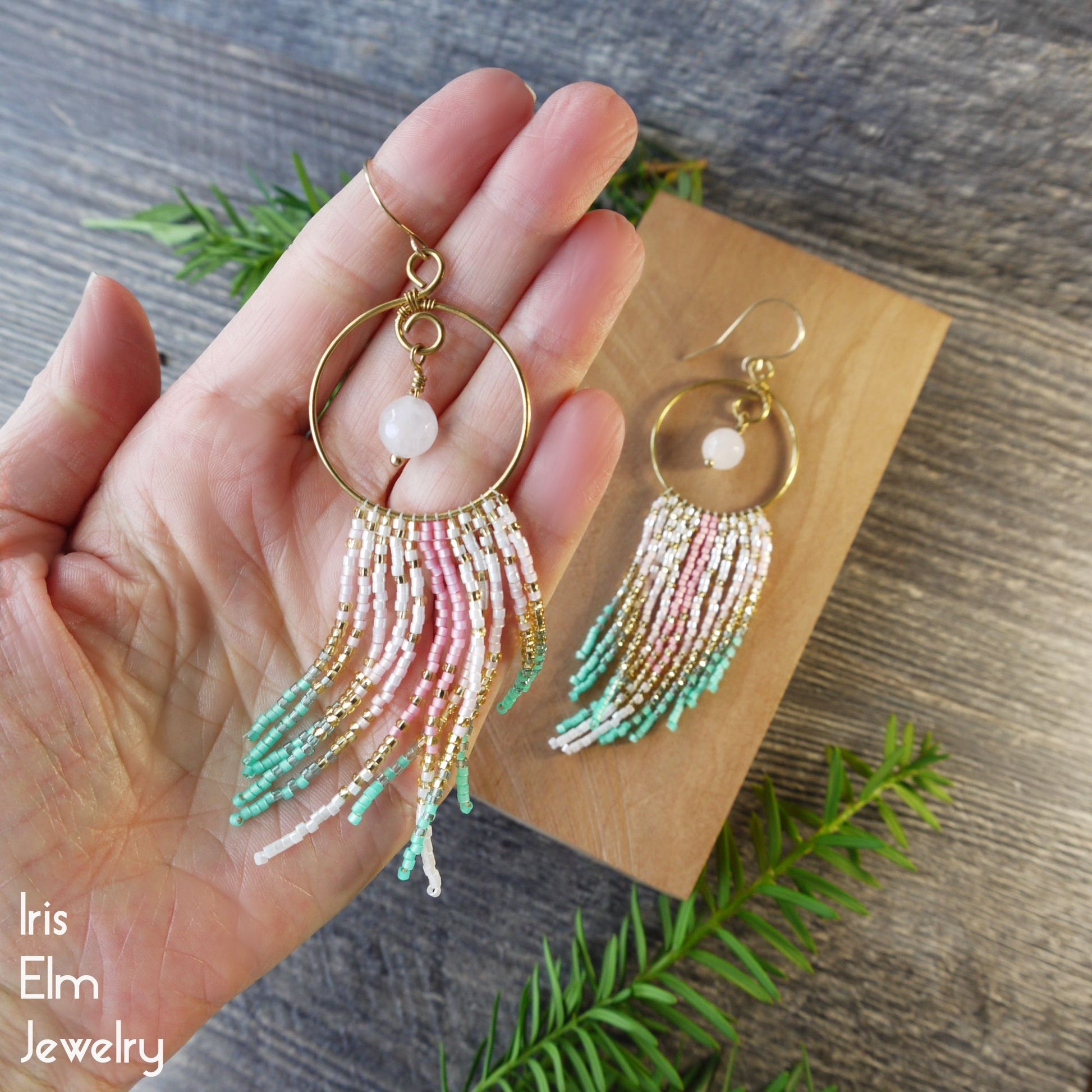 Gold hoop rose quartz earrings with colorful pink, gold, and mint green fringe held in a hand on a wooden surface.