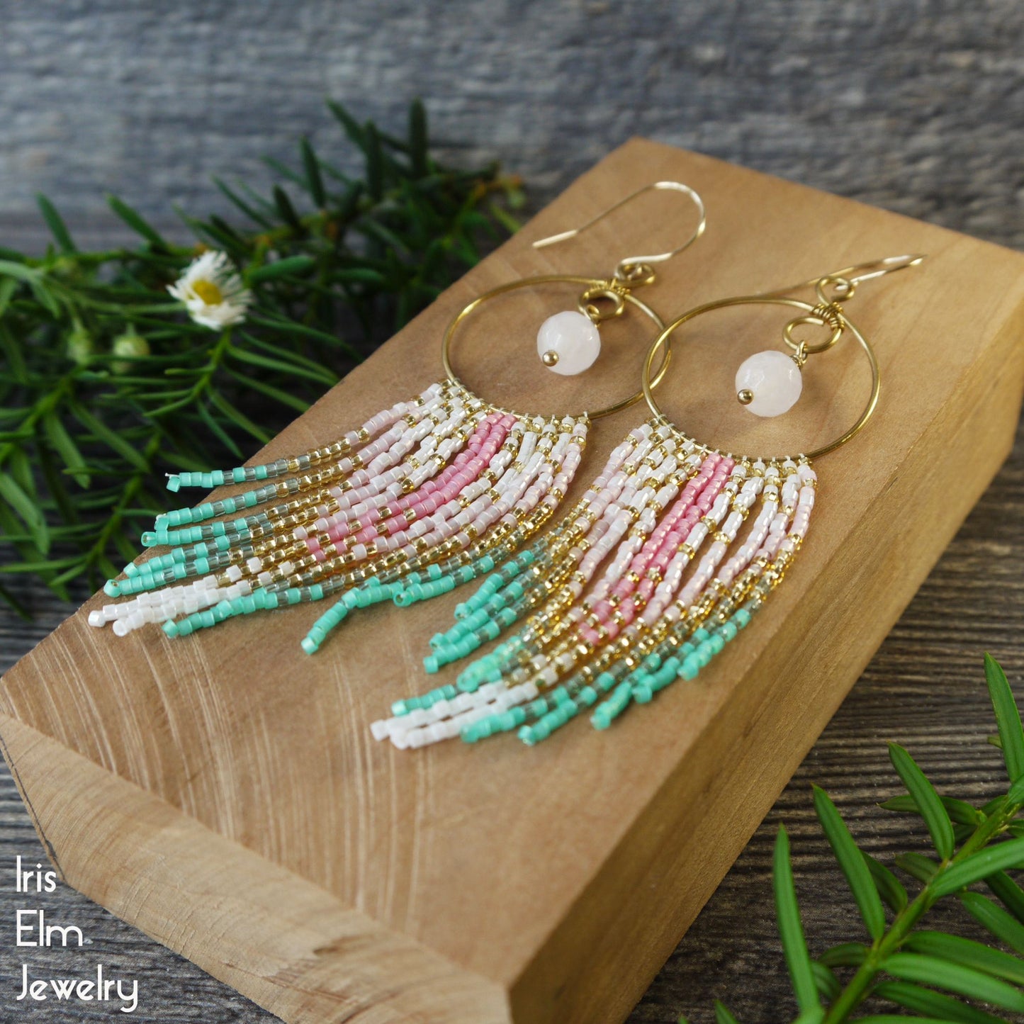 Beaded seed bead pink and green fringe hoop earrings with rose quartz gemstones on a wooden block with greenery in the background