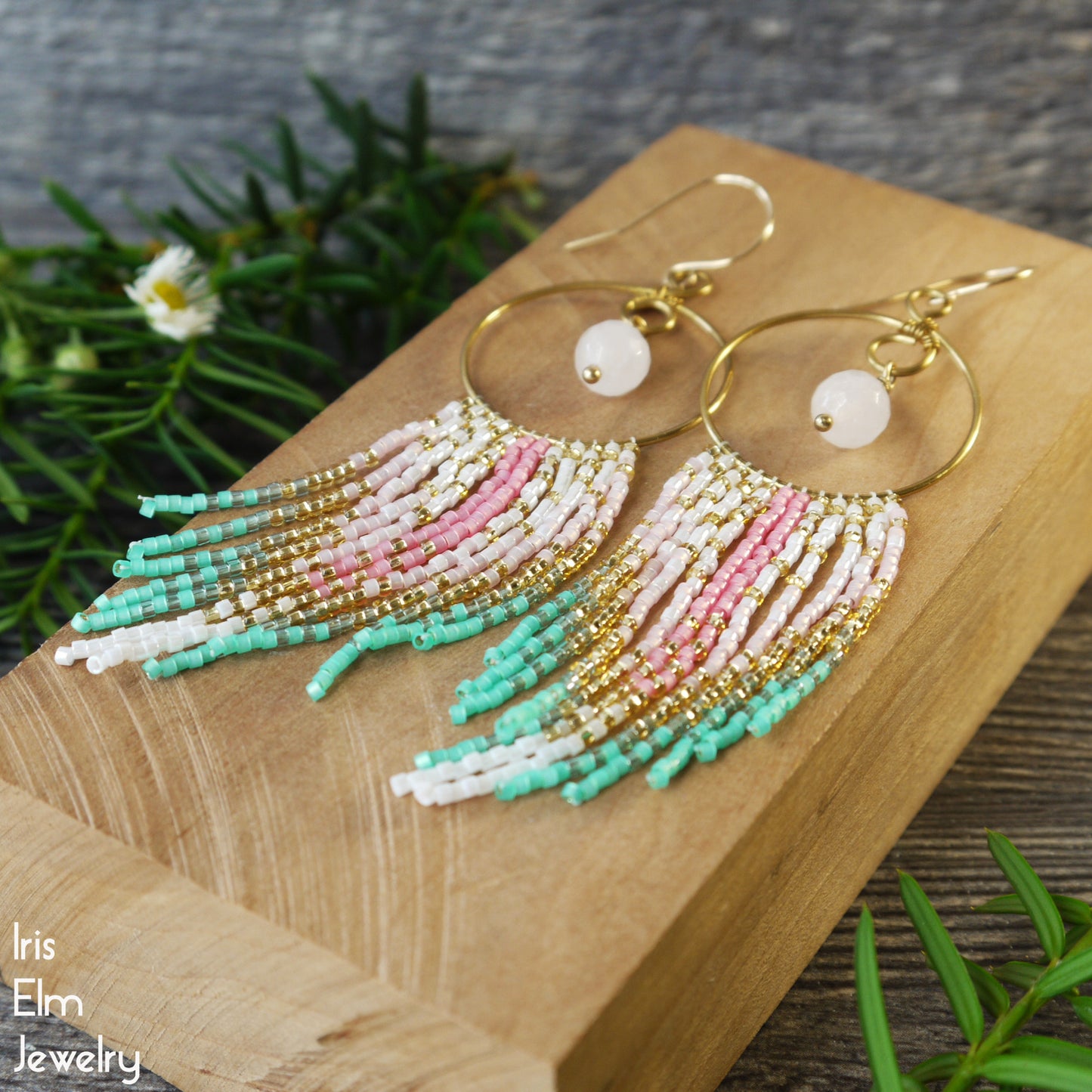 Beaded rose quartz hoop fringe earrings with pink and mint green beads on a wooden block with greenery in the background, featuring the brand 'Iris Elm Jewelry'.