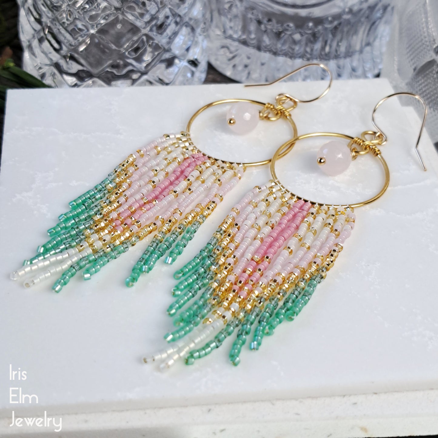 Beaded fringe handmade earrings with gold, pink, and green beads on a white surface with 'Iris Elm Jewelry' branding.