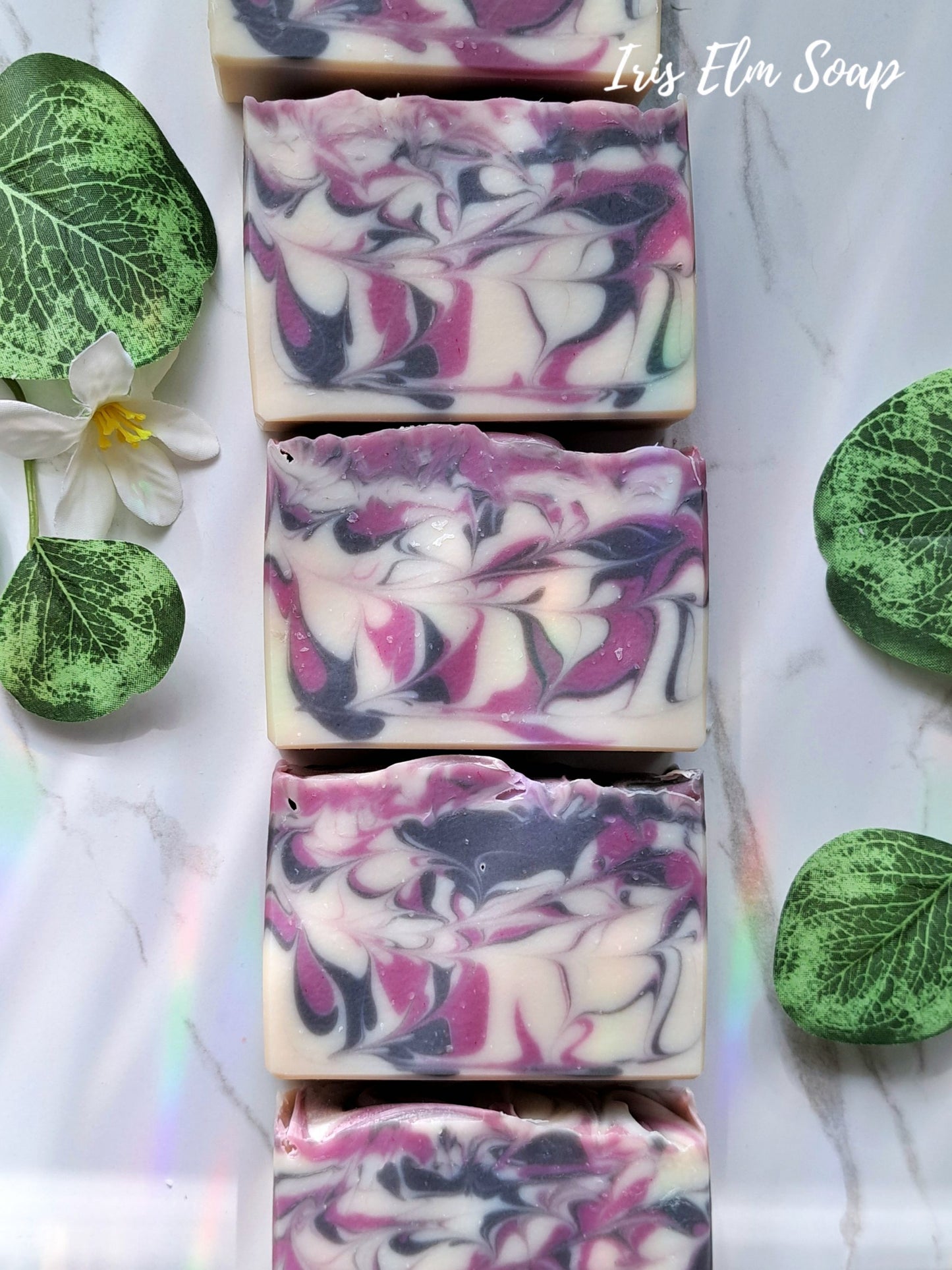Swirl-patterned black raspberry vanilla handmade soap bars with green leaves and a flower on a light background, branded 'Iris Elm Soap'.