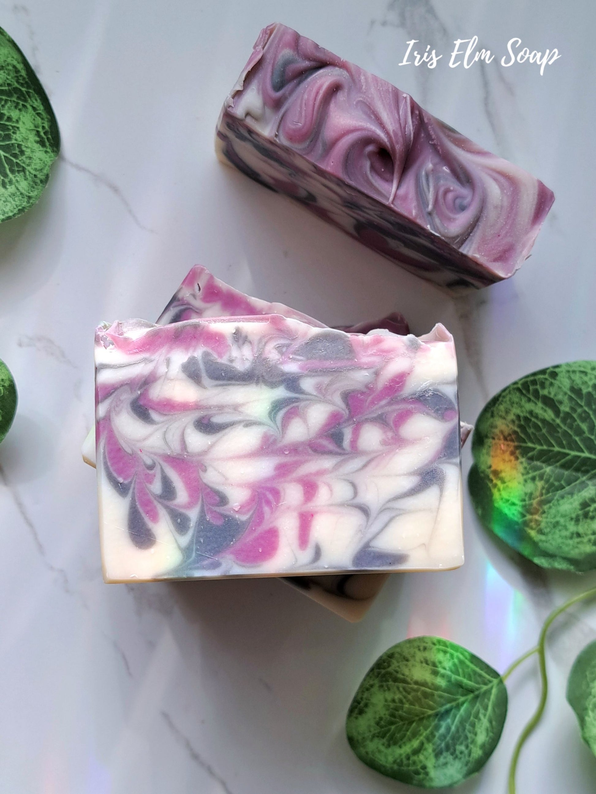 Two swirled black raspberry vanilla handmade soap bars on a white surface with green leaves.