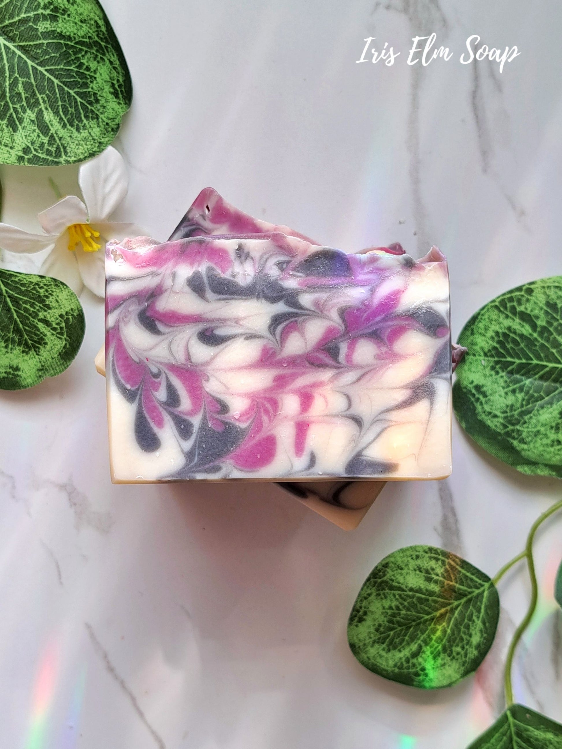 Swirled black raspberry vanilla handmade soap bar with pink and purple on a white background with green leaves