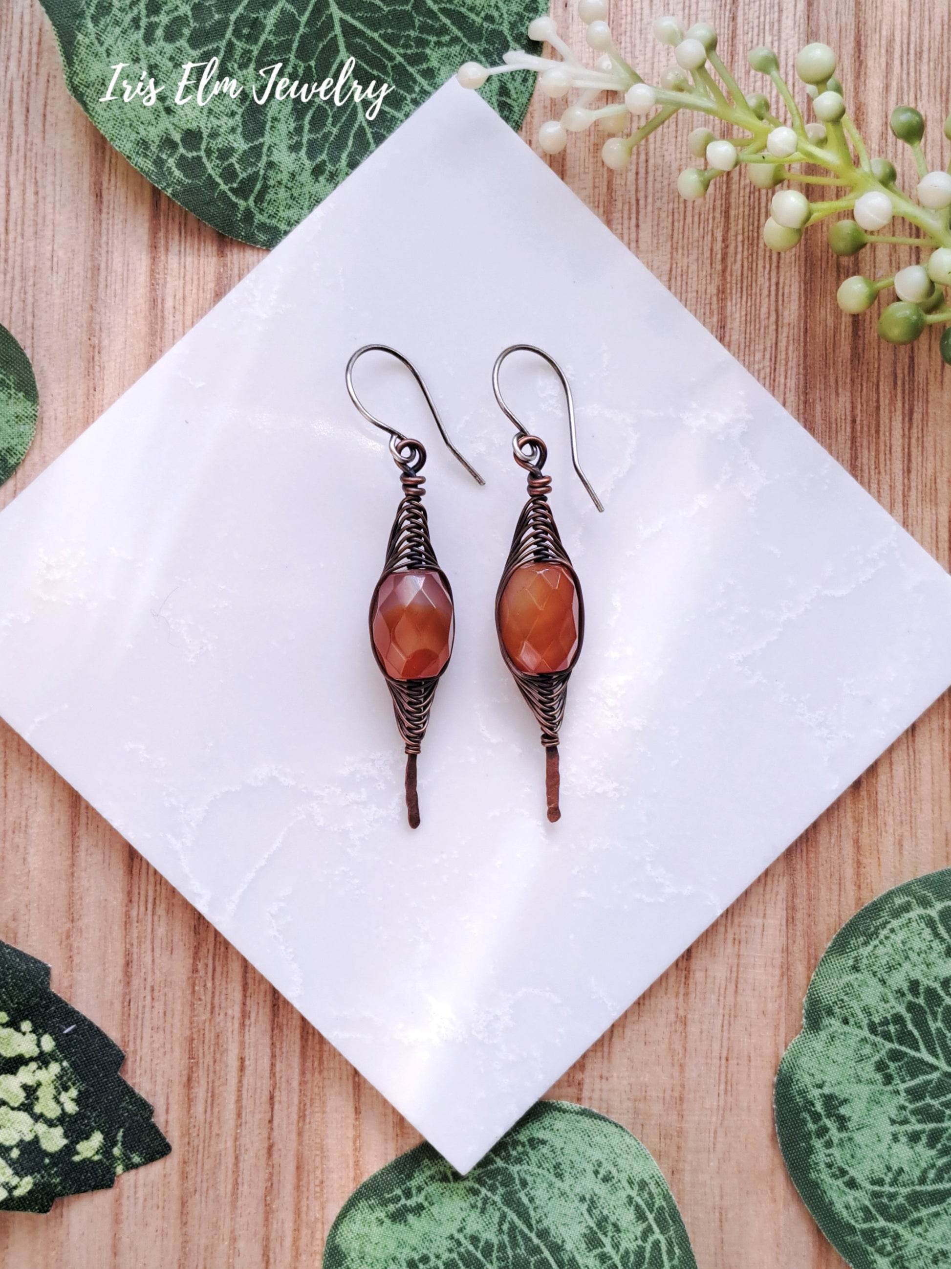 Pair of red agate copper wire wrapped herringbone earrings on a white tile with green leaves in the background