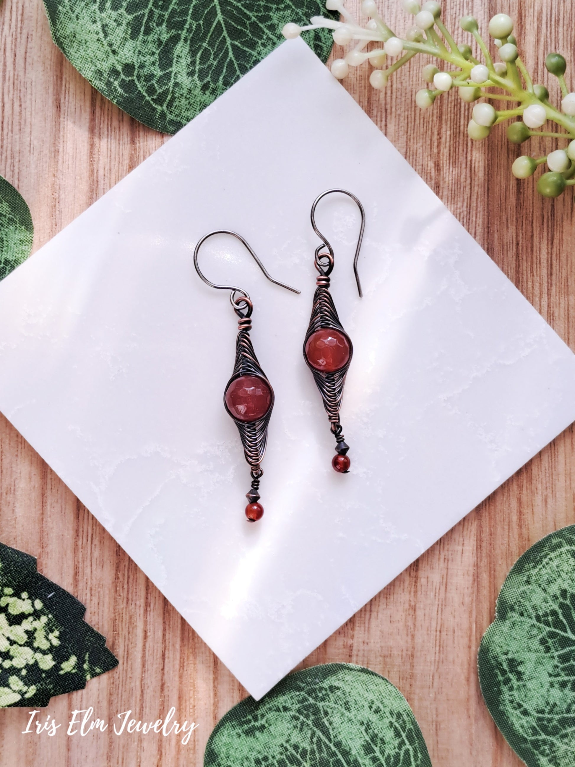 Pair of copper wire wrapped herringbone earrings with red agate stones on a white napkin with green leaves in the background