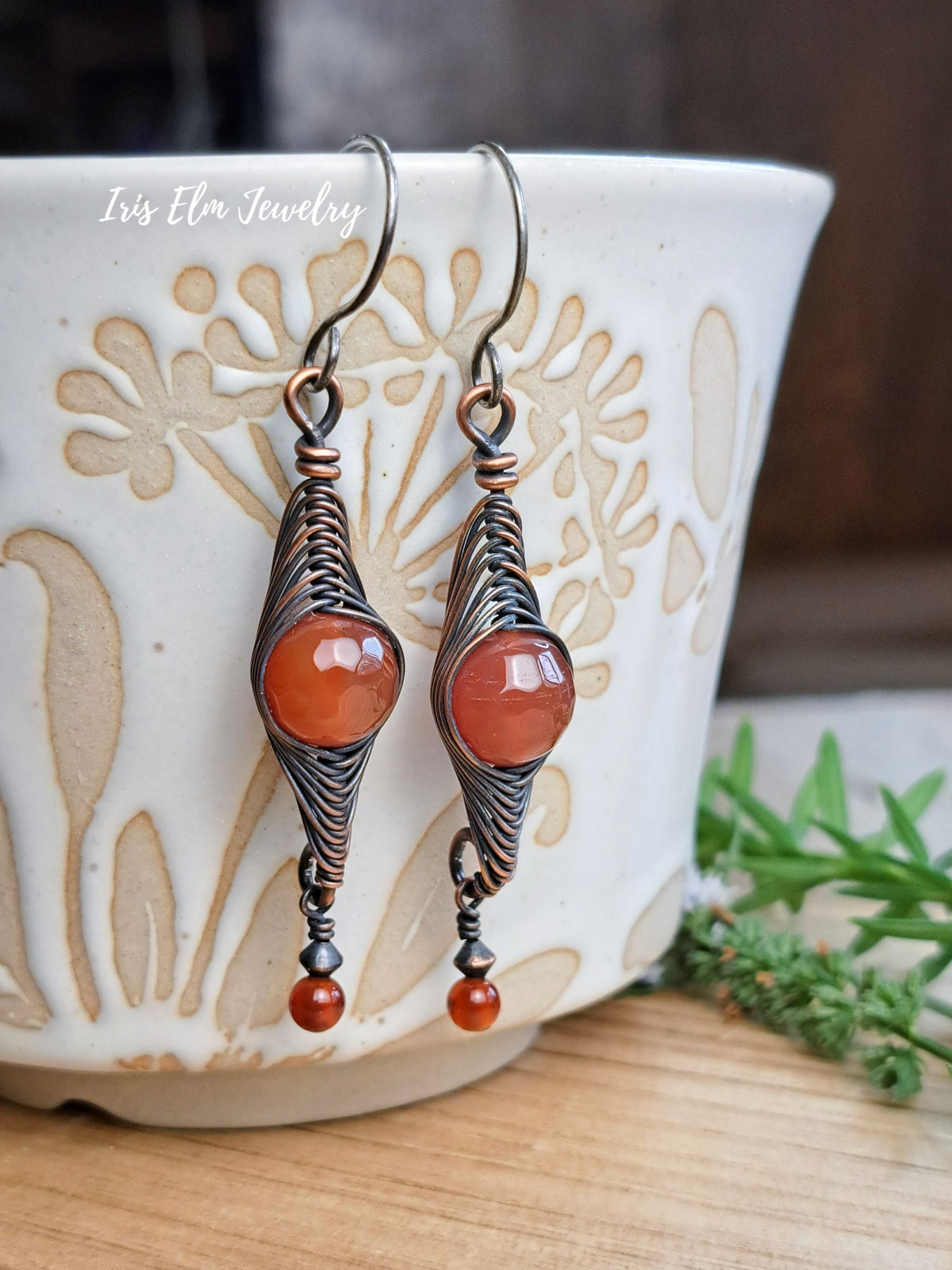 A pair of handmade dangle earrings by Iris Elm Jewelry, featuring copper wire wrapped herringbone pattern holding round orange cabochon stones, resting against a white ceramic cup on a wooden surface.