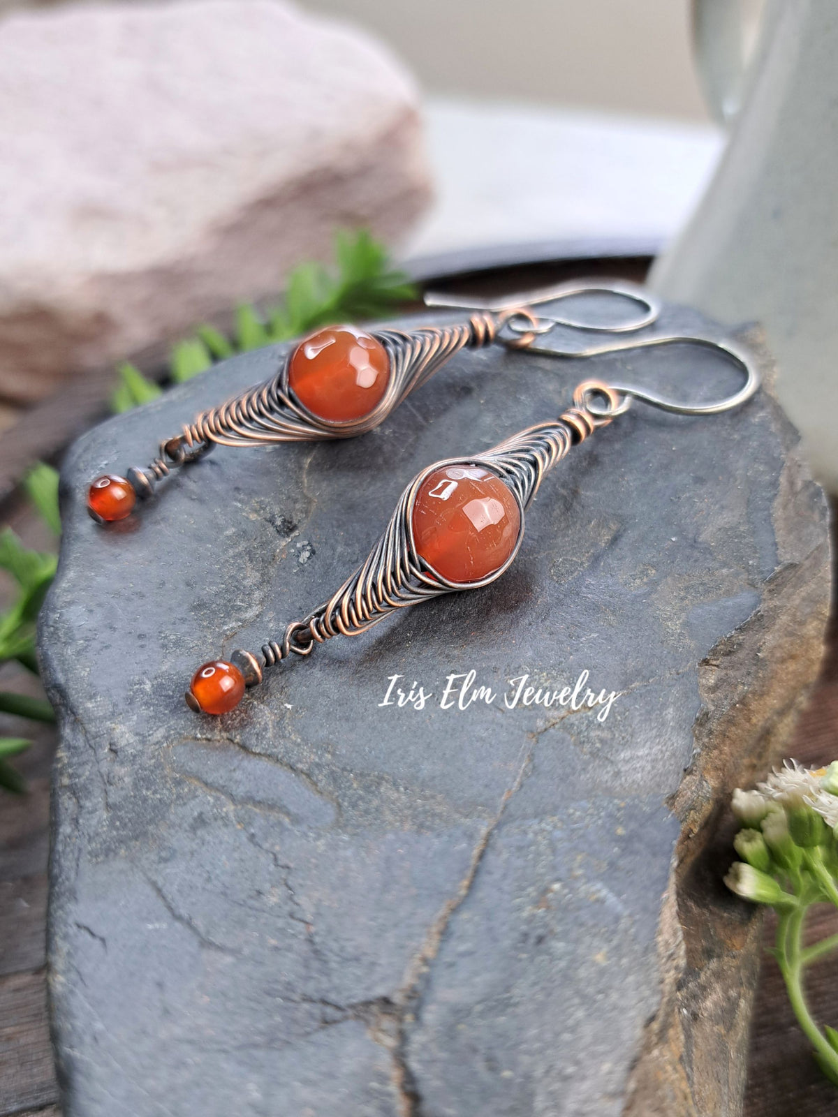 A pair of handmade dangle earrings with copper wire wrapping and orange agate stones, displayed on a dark slate stone with a soft-focus background.