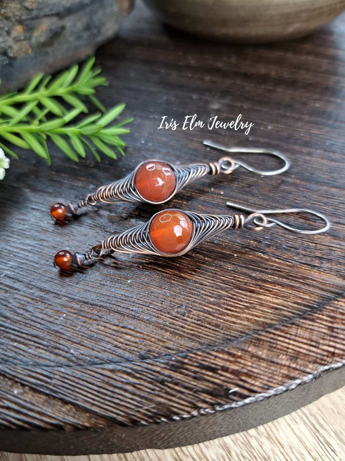 A pair of handmade dangle earrings with round red agate stone beads and silver-toned wire wrapping, displayed on a dark wooden surface with a green sprig and a ceramic bowl in the background. The text 'Iris Elm Jewelry' is visible on the image.