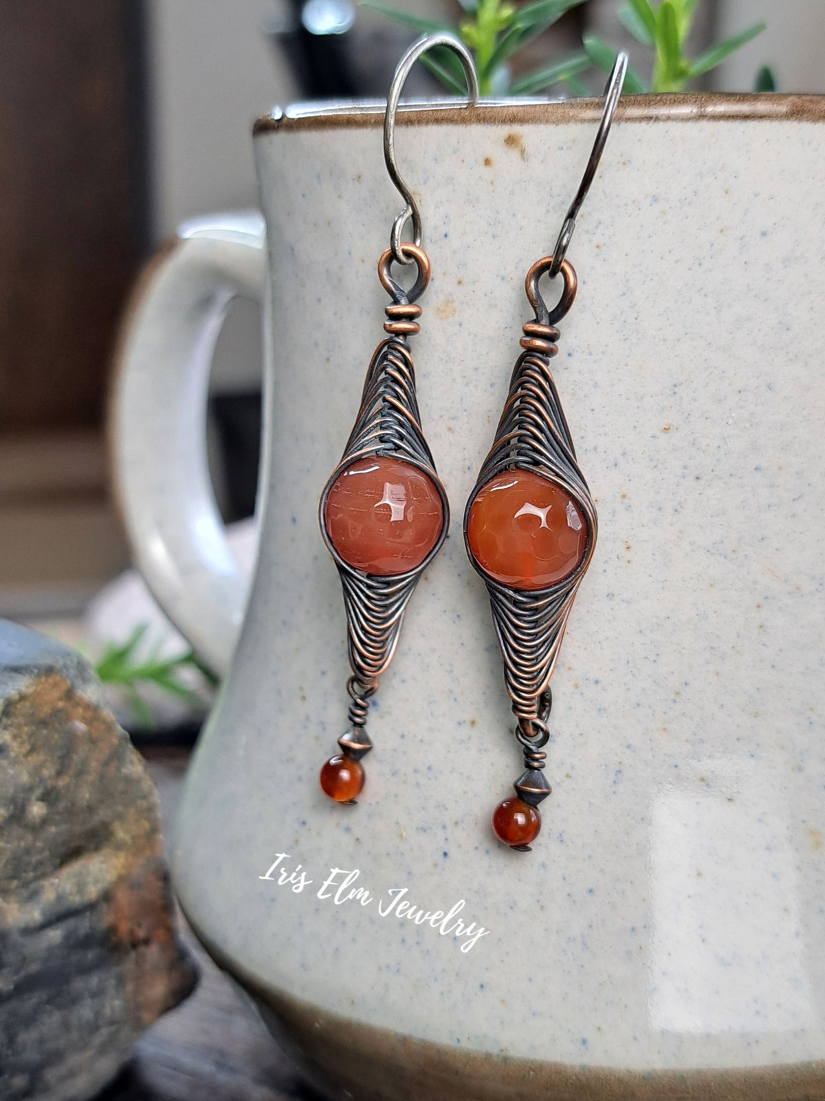 A pair of handmade dangle earrings with copper wire wrapped details and round orange cabochon gemstones, hanging in front of a speckled ceramic mug. The brand name 'Iris Elm Jewelry' is visible as a watermark on the mug.