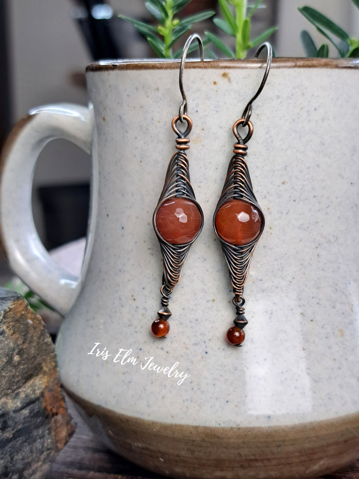 A pair of handmade dangle earrings with copper wire wrapped teardrop shapes and round red agate gemstone centers, hanging in front of a speckled ceramic mug. The text 'Iris Elm Jewelry' is visible on the mug.