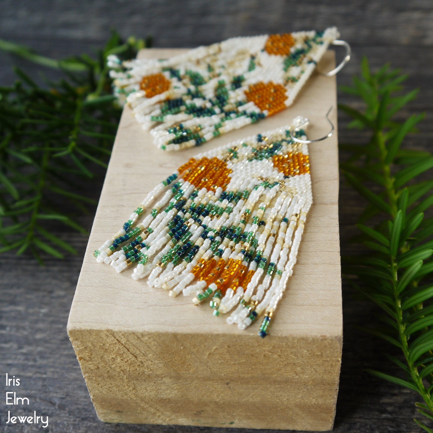 Beaded fringe seed bead earrings  with orange floral design on a wooden block with greenery in the background, featuring the brand 'Iris Elm Jewelry'.