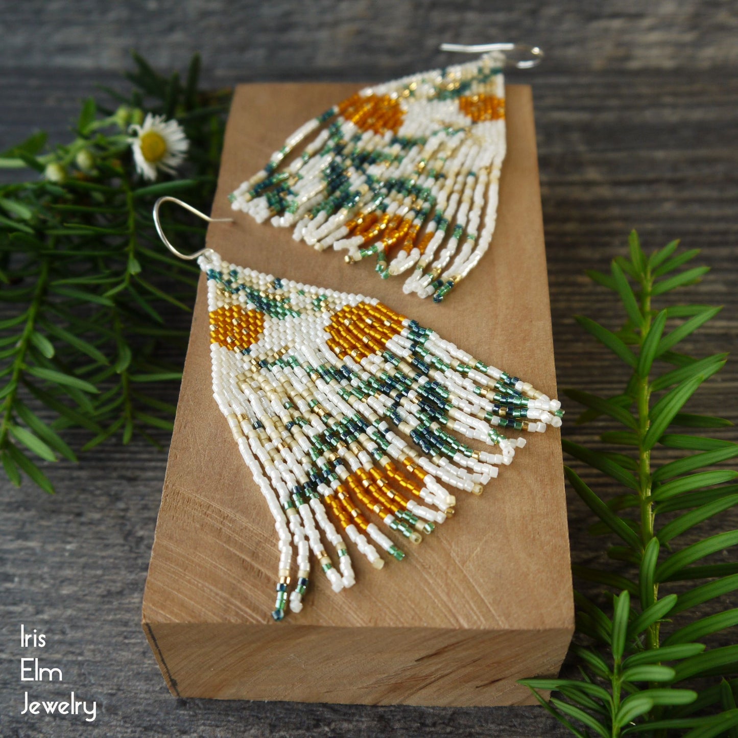Beaded orange floral fringe handmade beadwoven earrings on a wooden block with greenery in the background, featuring the brand 'Iris Elm Jewelry'.