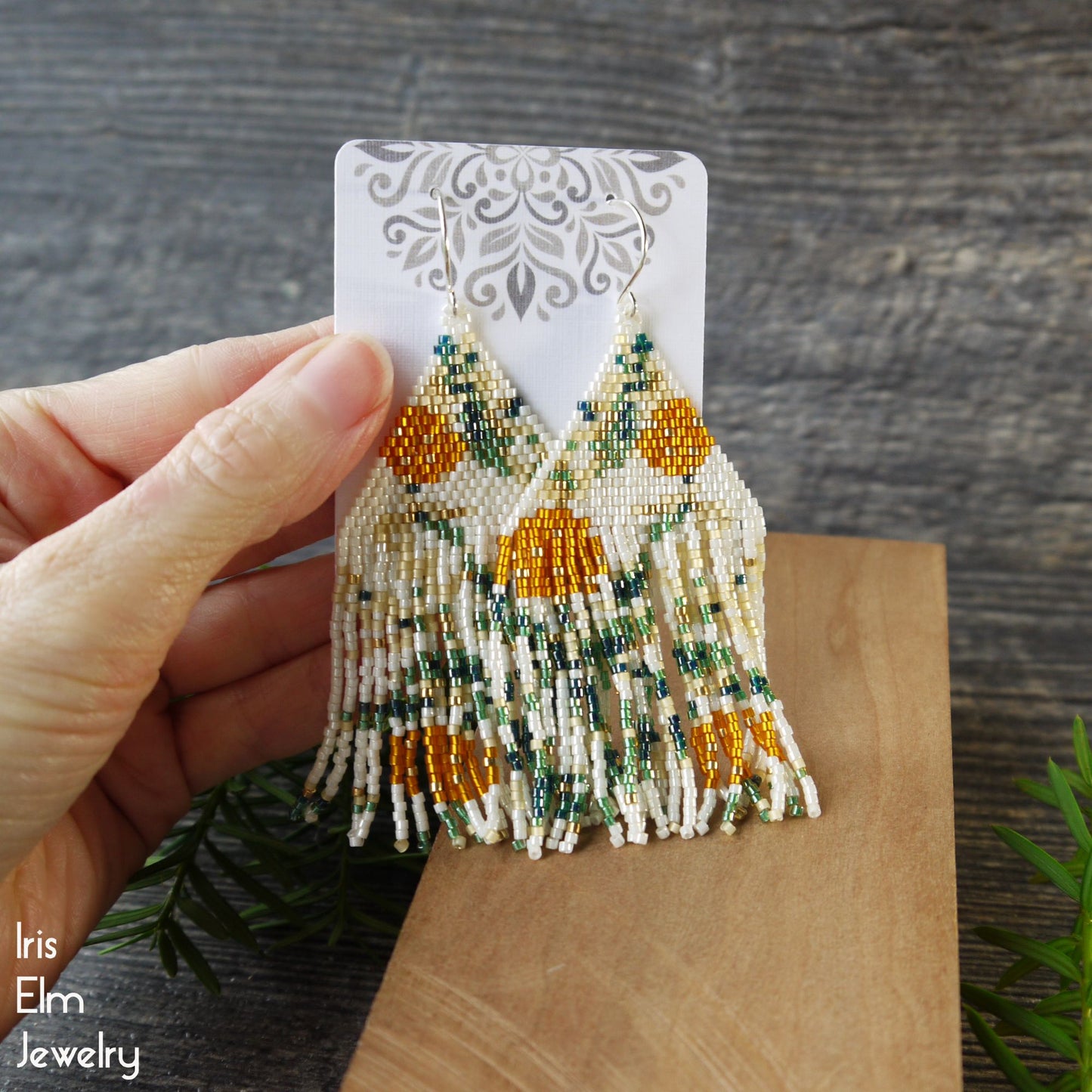 Beaded seed bead orange flower fringe earrings held by a hand on a wooden surface with 'Iris Elm Jewelry' branding.