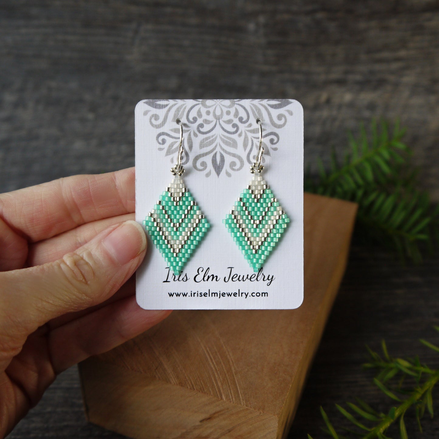 Green beaded earrings on a 'Iris Elm Jewelry' card held by a hand with a wooden surface and greenery in the background.