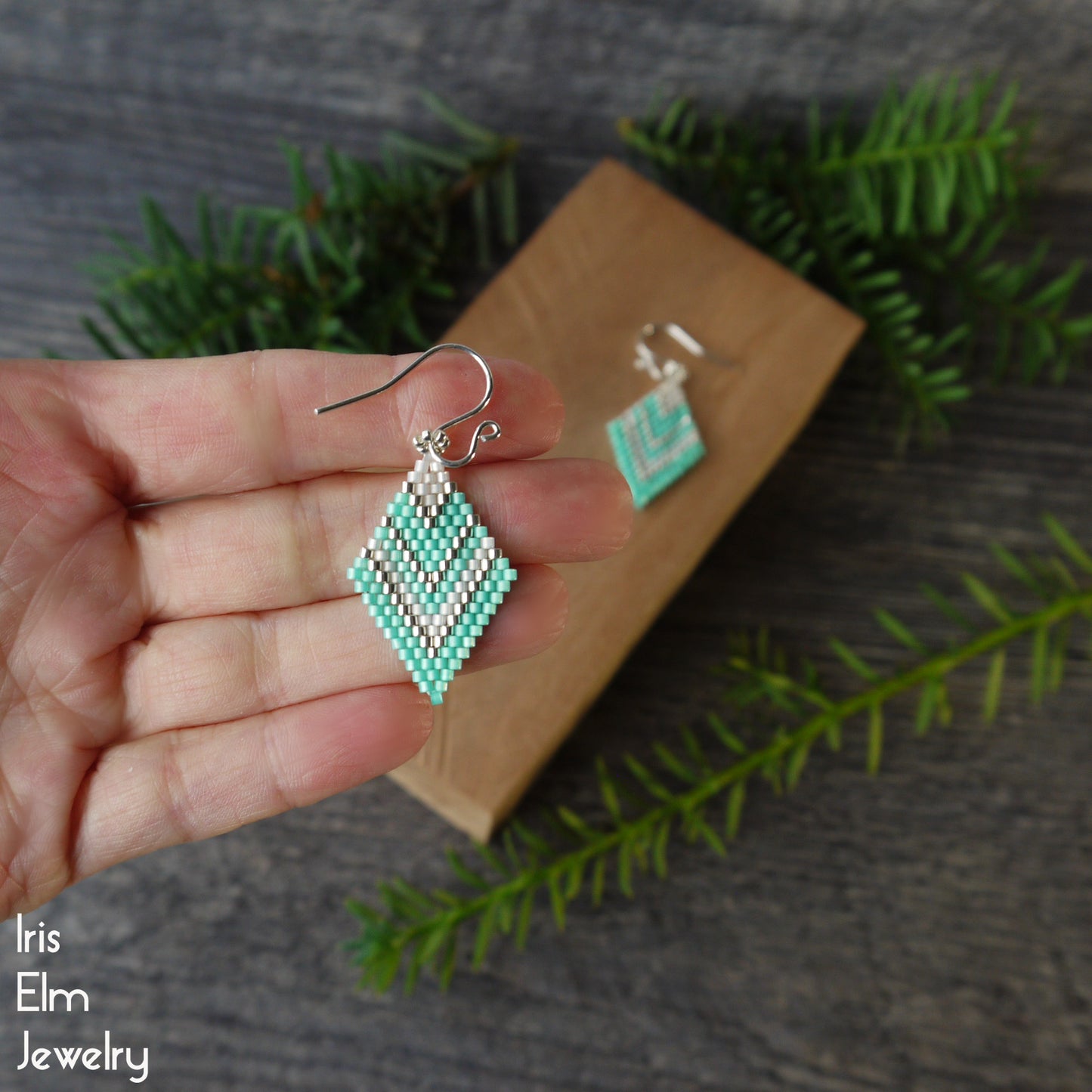 mint green diamond shaped beaded earrings held in a hand with a wooden box and green leaves in the background, featuring 'Iris Elm Jewelry'.