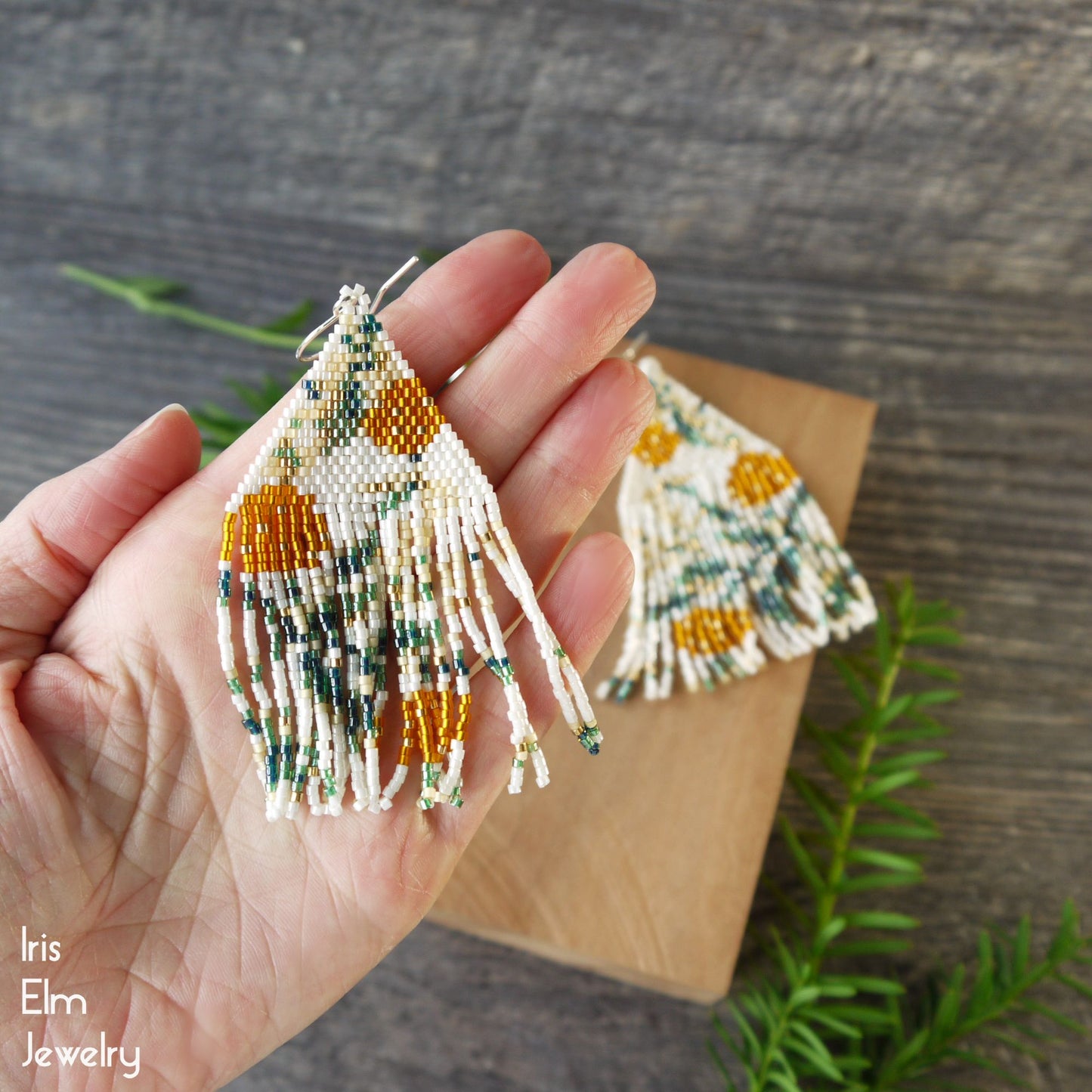 Hand holding beaded orange floral and cream seed bead fringe earrings with a wooden block and green leaves in the background