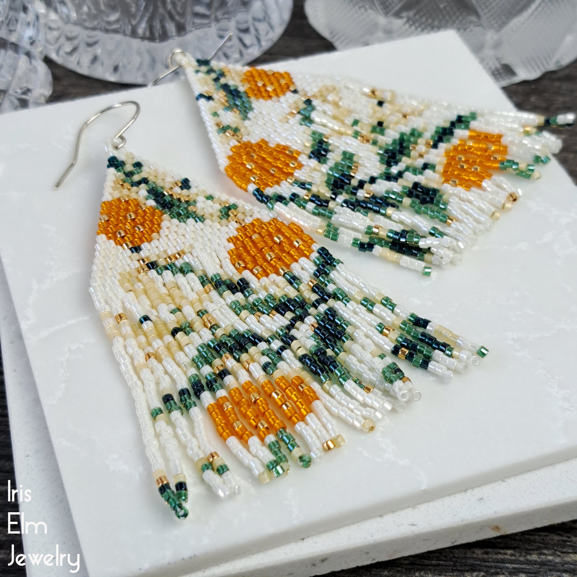 Beaded fringe statement earrings with orange and green floral and leaf patterns on a marble surface, featuring 'Iris Elm Jewelry' branding.