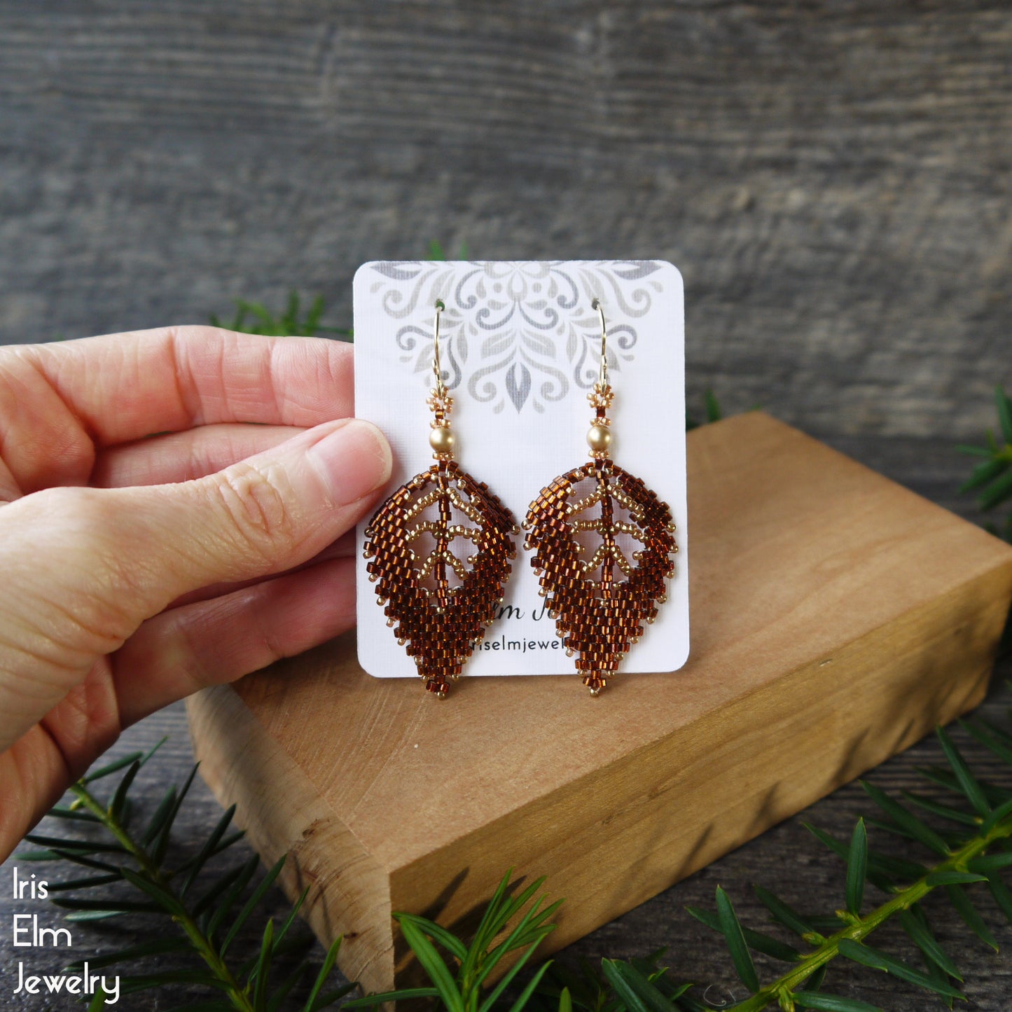Brown beaded leaf handmade earrings on a card held by a hand with a wooden block and greenery in the background.