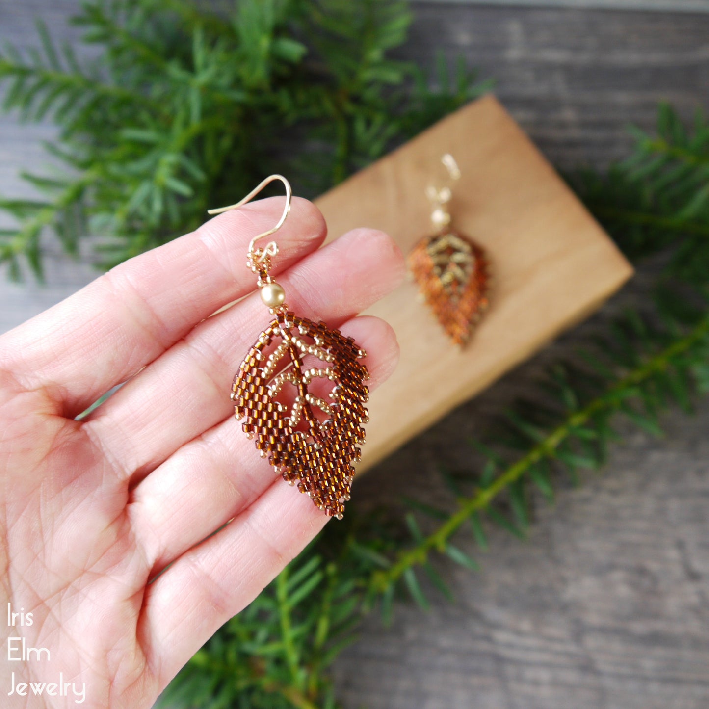 Gold and topaz brown handmade leaf-shaped earrings held in a hand with a wooden box and greenery in the background.