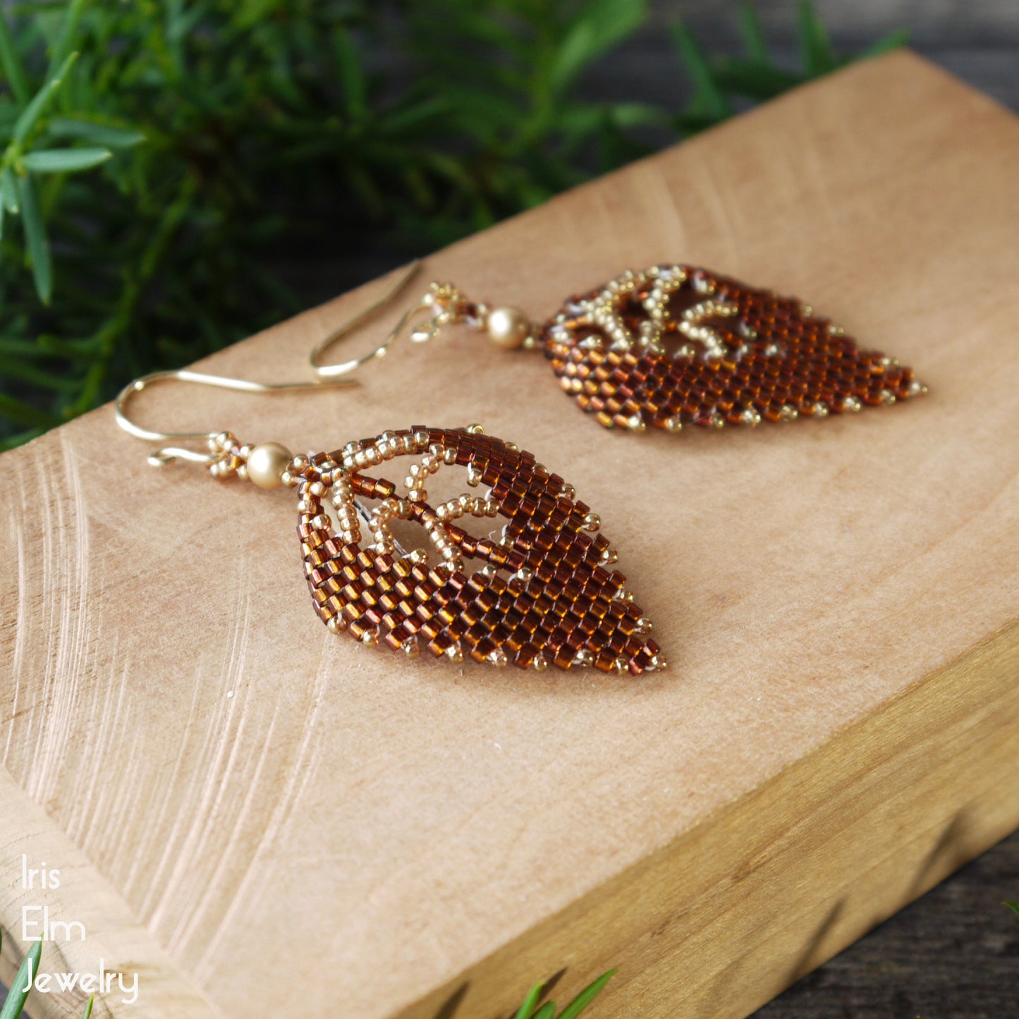 Gold and brown leaf-shaped earrings on a wooden block with greenery in the background