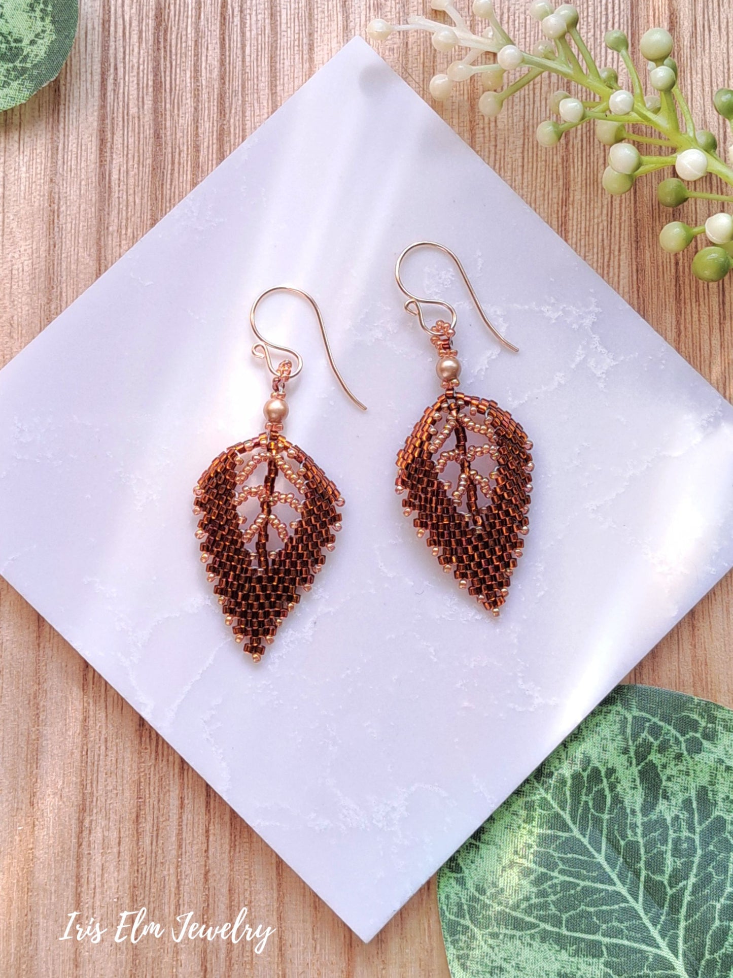 Pair of leaf-shaped autumn seed bead brown earrings on a white napkin with green leaves in the background